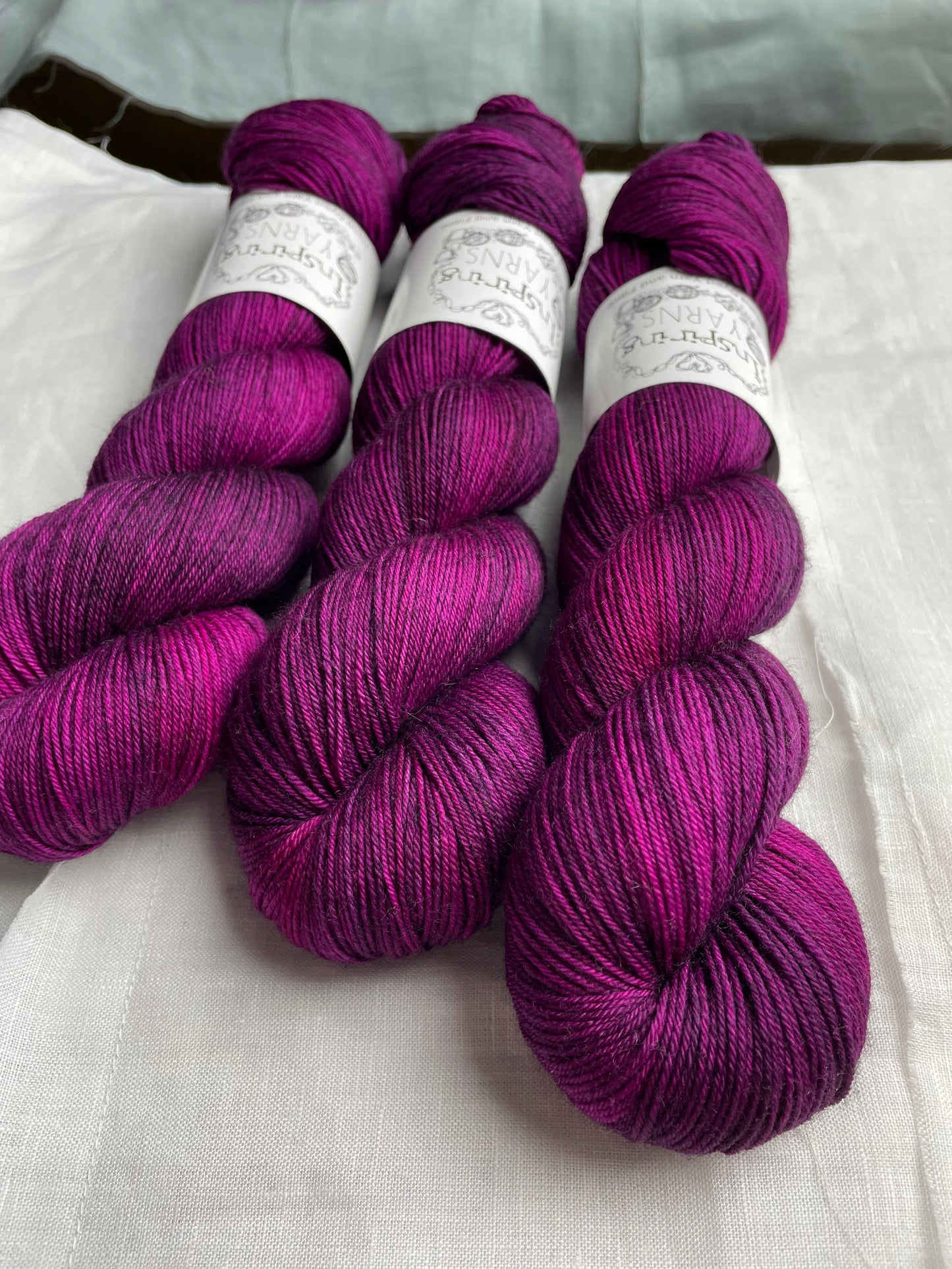 Three lightly variegated cerise pink and dark purple coloured skeins of Merino Smooth Sock yarn branded Inspiring Yarns on a white linen fabric background.