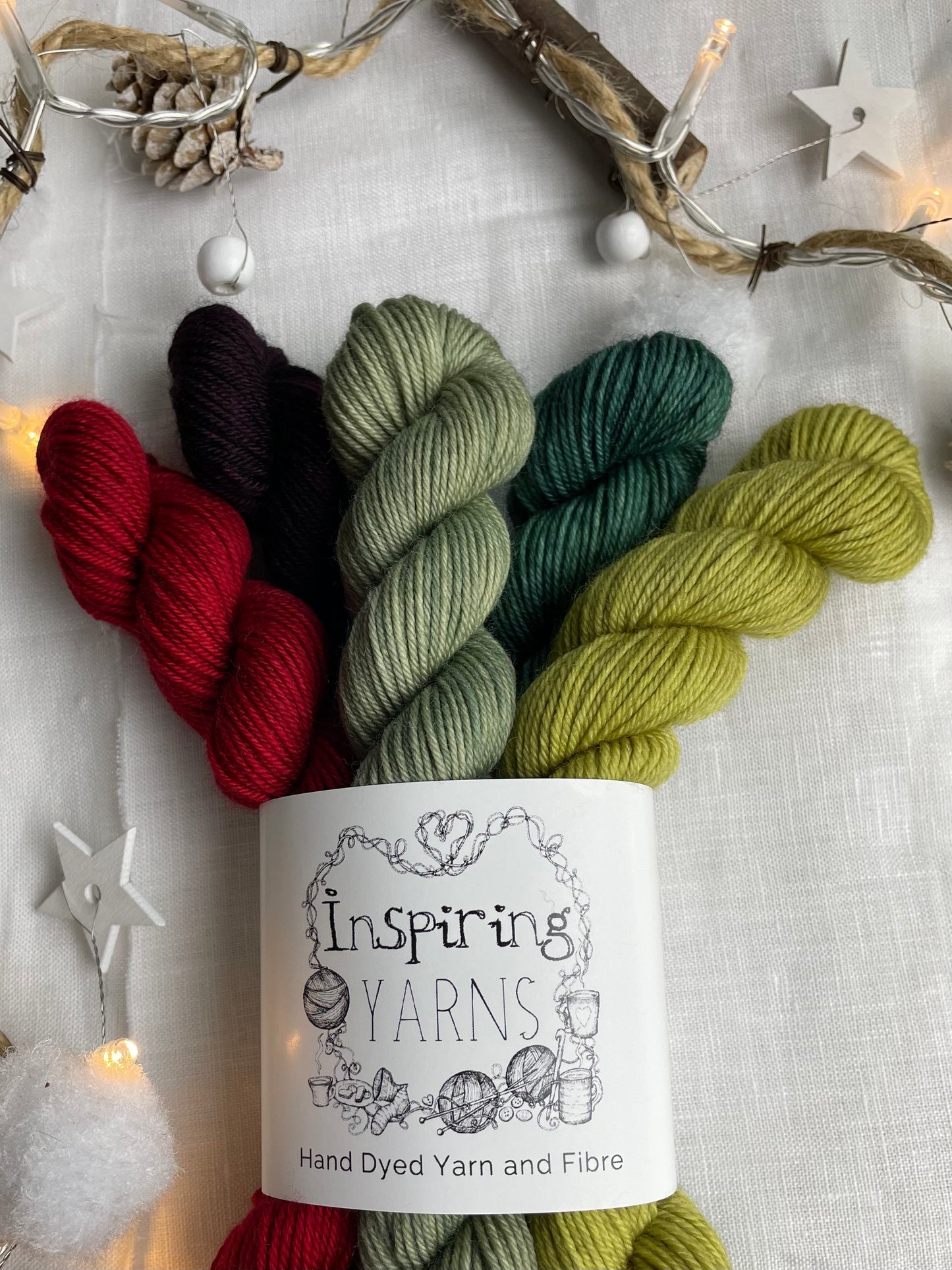 A set of five hand dyed yarn mini skeins on Merino Smooth Sock Yarn in red and green tones labelled Inspiring Yarns. The background is white linen and some Christmas lights with pinecones, twigs and stars.