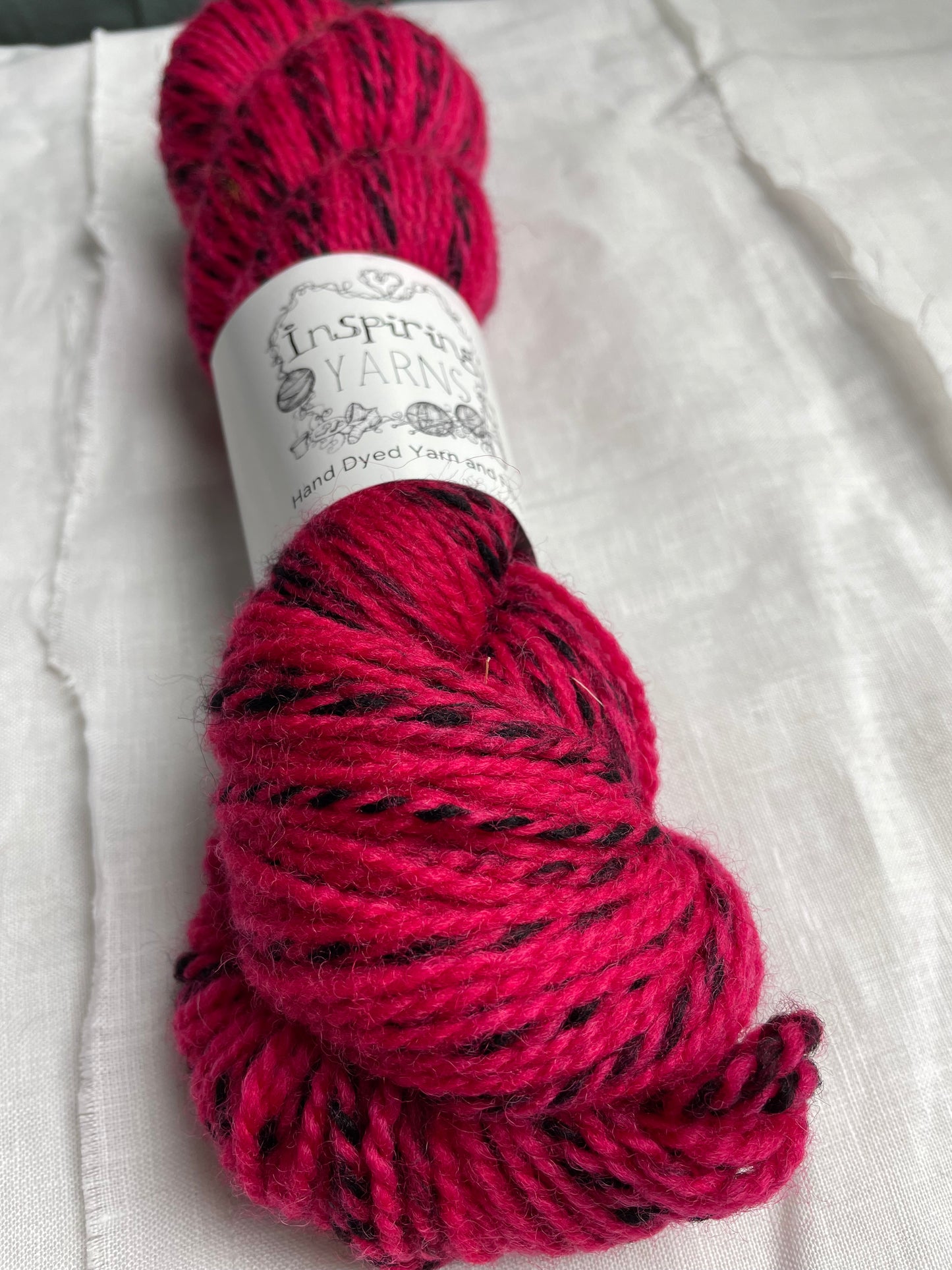 Close up of oneskein of handyed Highland Zebra yarn in a bright cerise pink colourway. The yarn has black strands spun in to it. the skein is labelled as Inspiring Yarns.