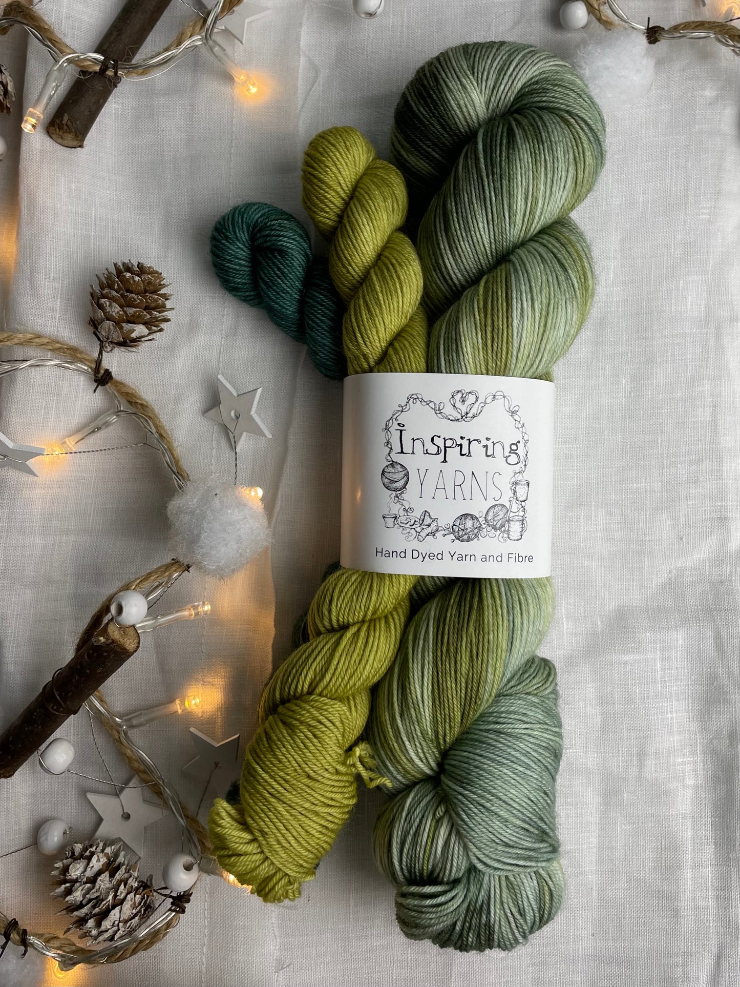 A hand dyed yarn sock set featuring large skein in green tones with a teal and a lime green mini skein labelled Inspiring Yarns. The background is white linen and some Christmas lights with pinecones, twigs and stars.