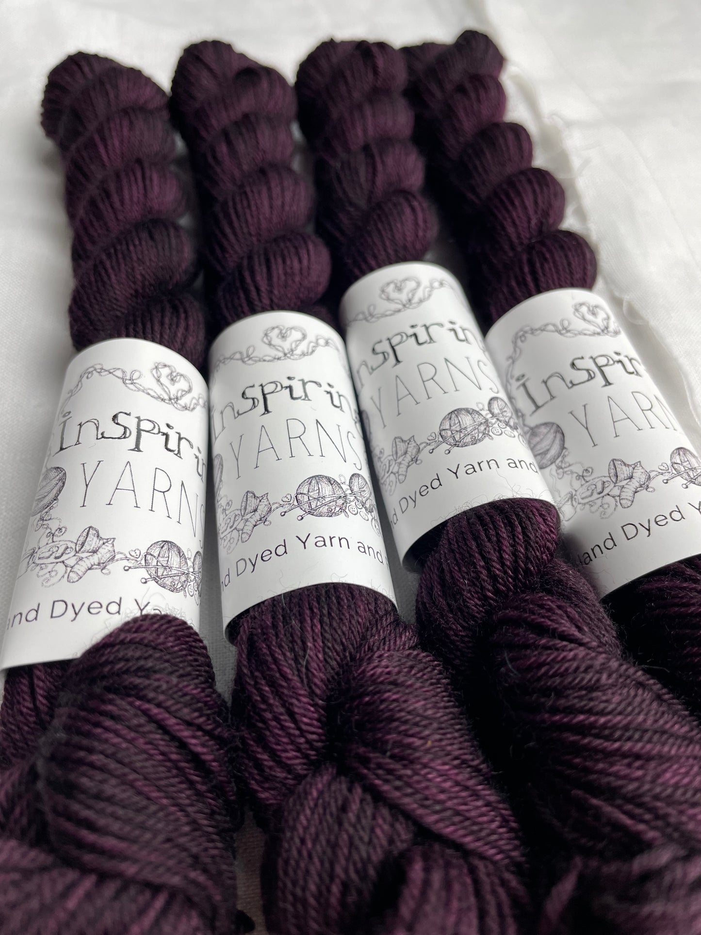 Four skeins of merino smooth sock yarn in a tonal dark burgundy colour labelled Inspiring Yarns on a white linen background.