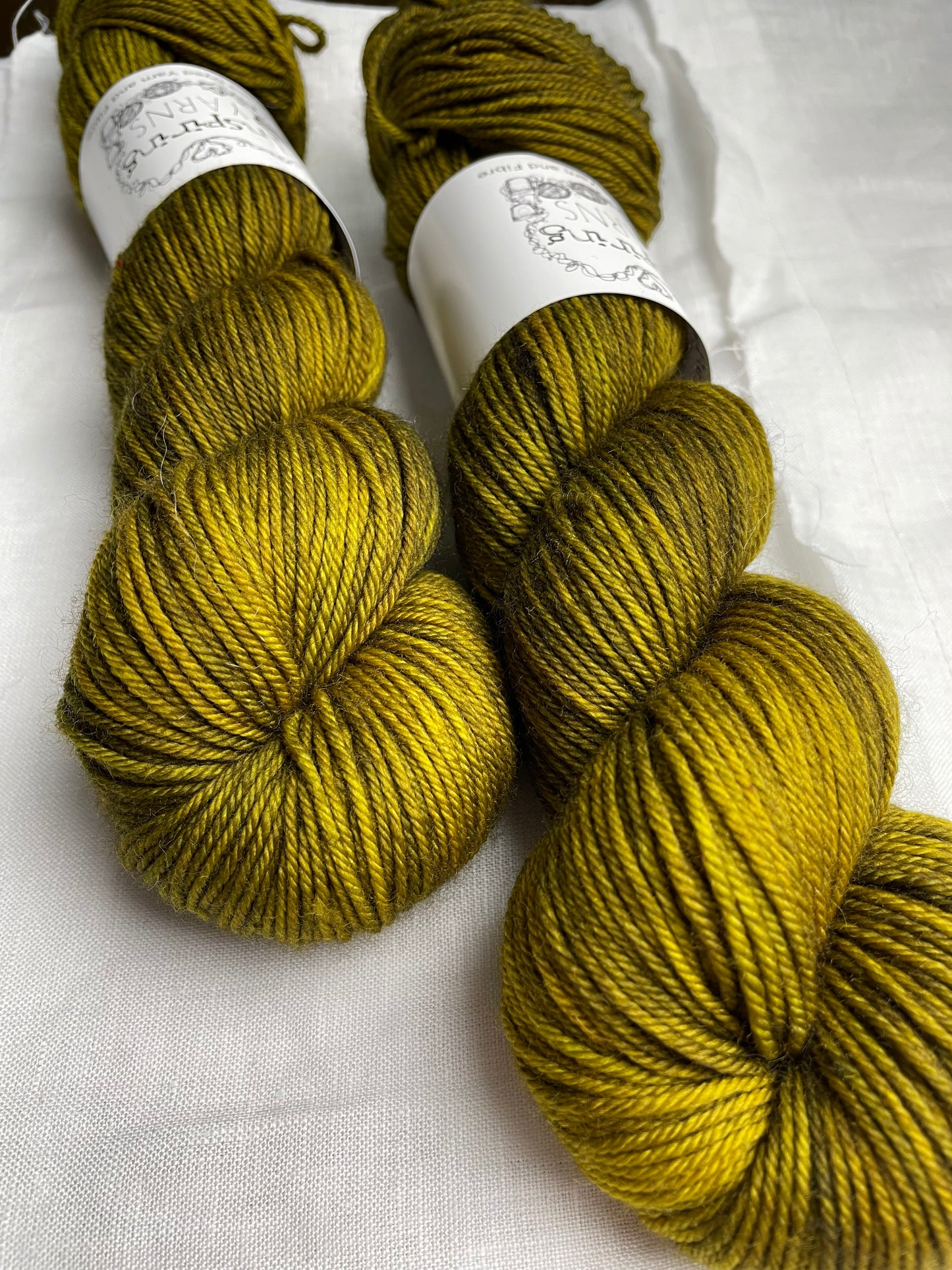 Close up of two skeins of dark olive green Merino Smooth DK yarn labelled Inspiring Yarns on a white linen background.