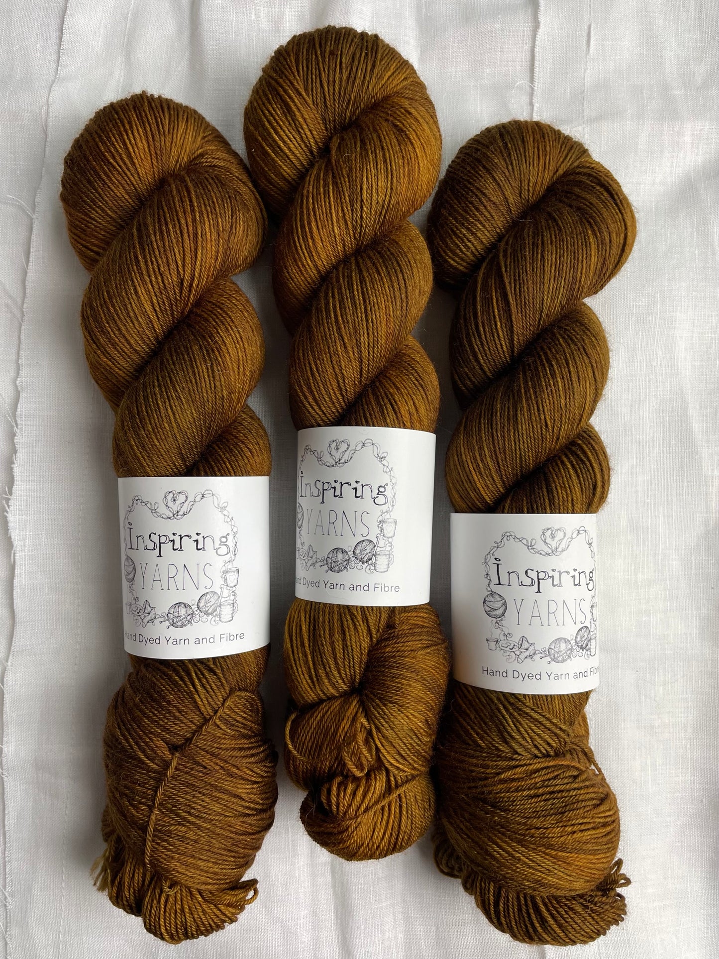 Three skeins of Kelp, a bronze coloured Merino Smooth Sock, labelled Inspiring Yarns on a white linen background.