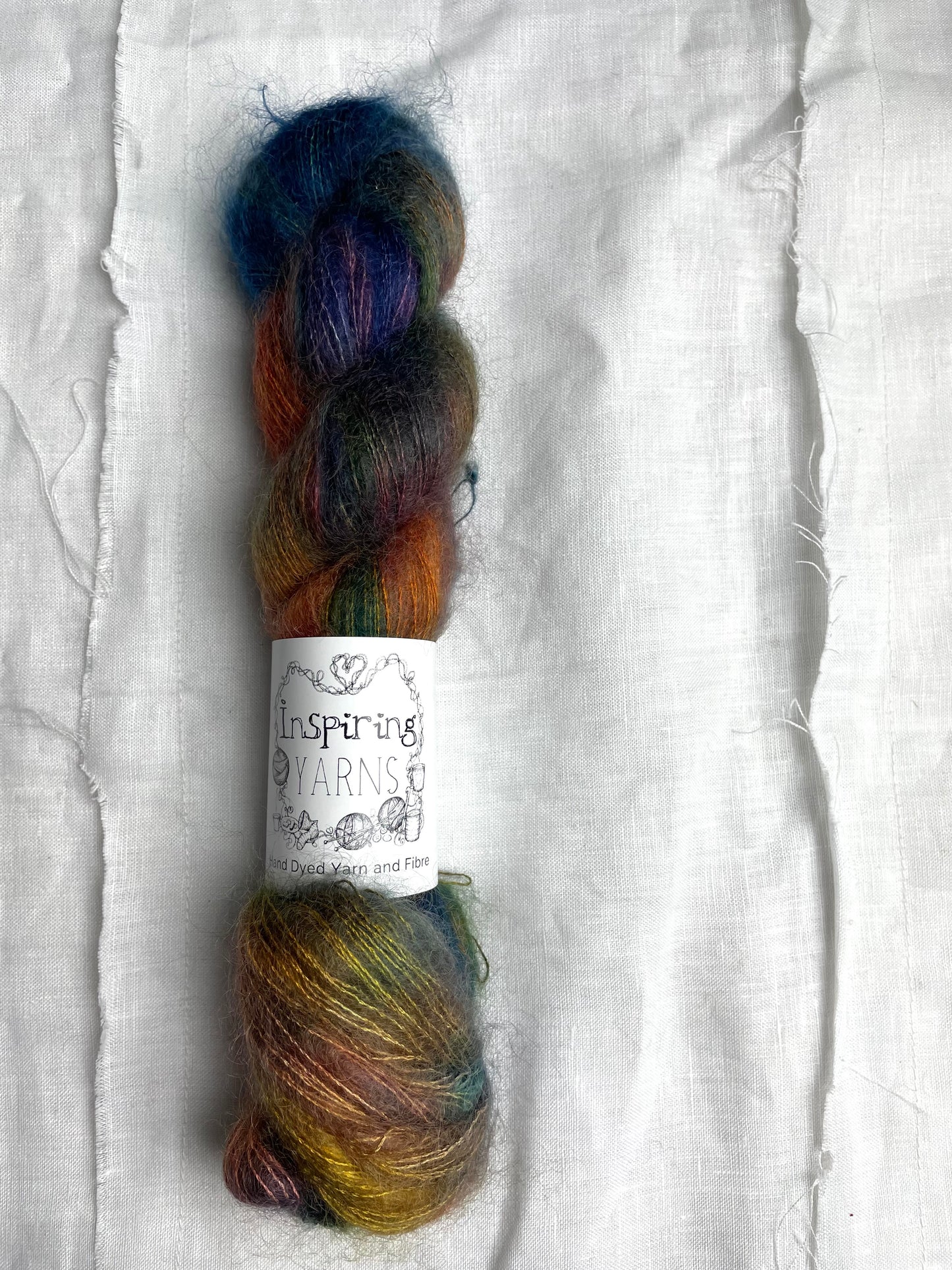 A skein of variegated blue and multi coloured kid mohair silk lace yarn labelled Inspiring Yarns on a white linen background.