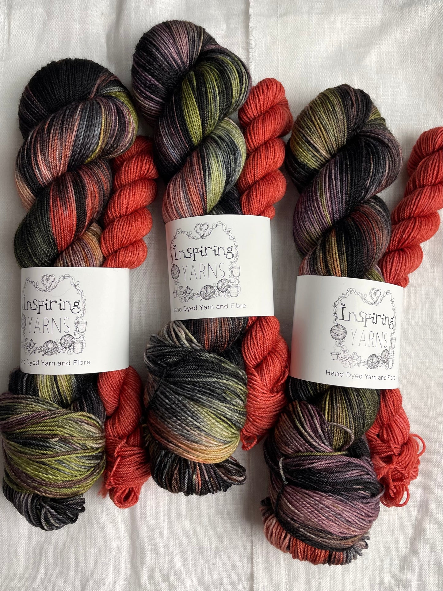 three sock sets made of a skein of black and multi coloured hand dyed yarn with a contrasting deep terraccotta mini skein, labelled Inspiring Yarns and laid on a white linen cloth.