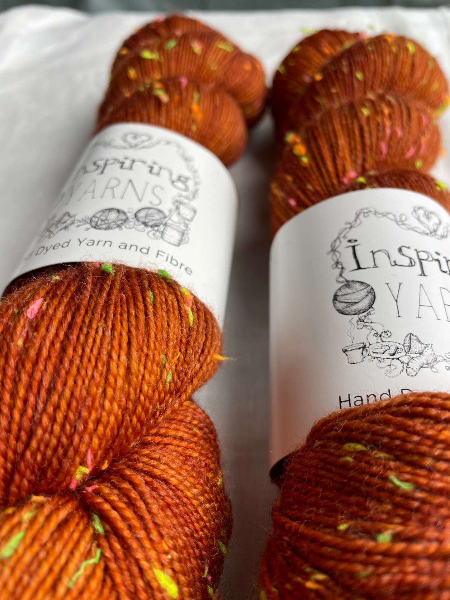 Close up of two skeins of tonal burnt orange coloured hand dyed Disco Donegal yarn branded Inspiring Yarns on a white linen background. The yarn has multi coloured neon neps.