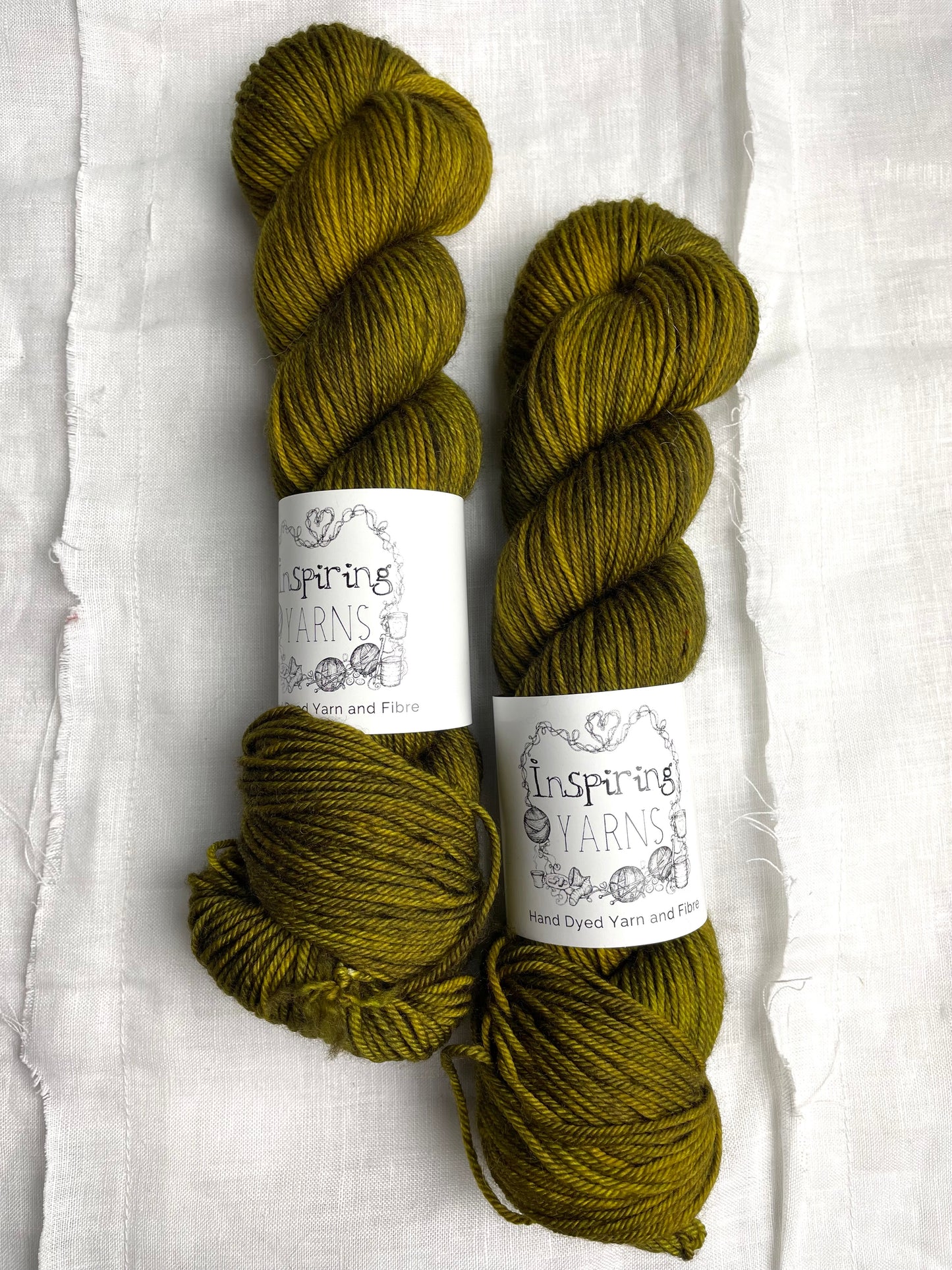 Two skeins of dark olive green Merino Smooth DK yarn labelled Inspiring Yarns on a white linen background.