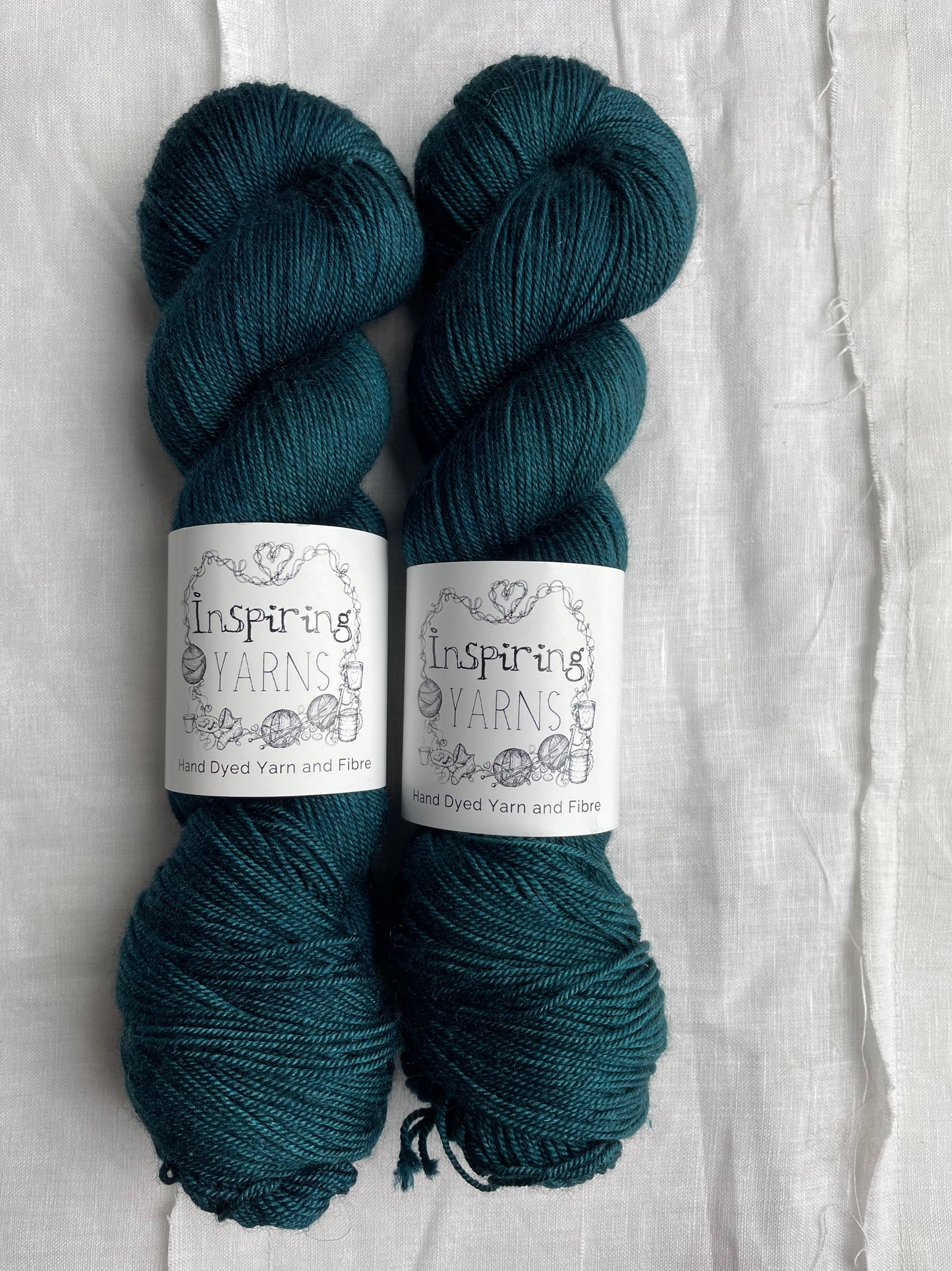 Two tonal intense teal blue coloured skeins of hand dyed Yak Silk 4Ply yarn branded Inspiring Yarns on a white linen background.