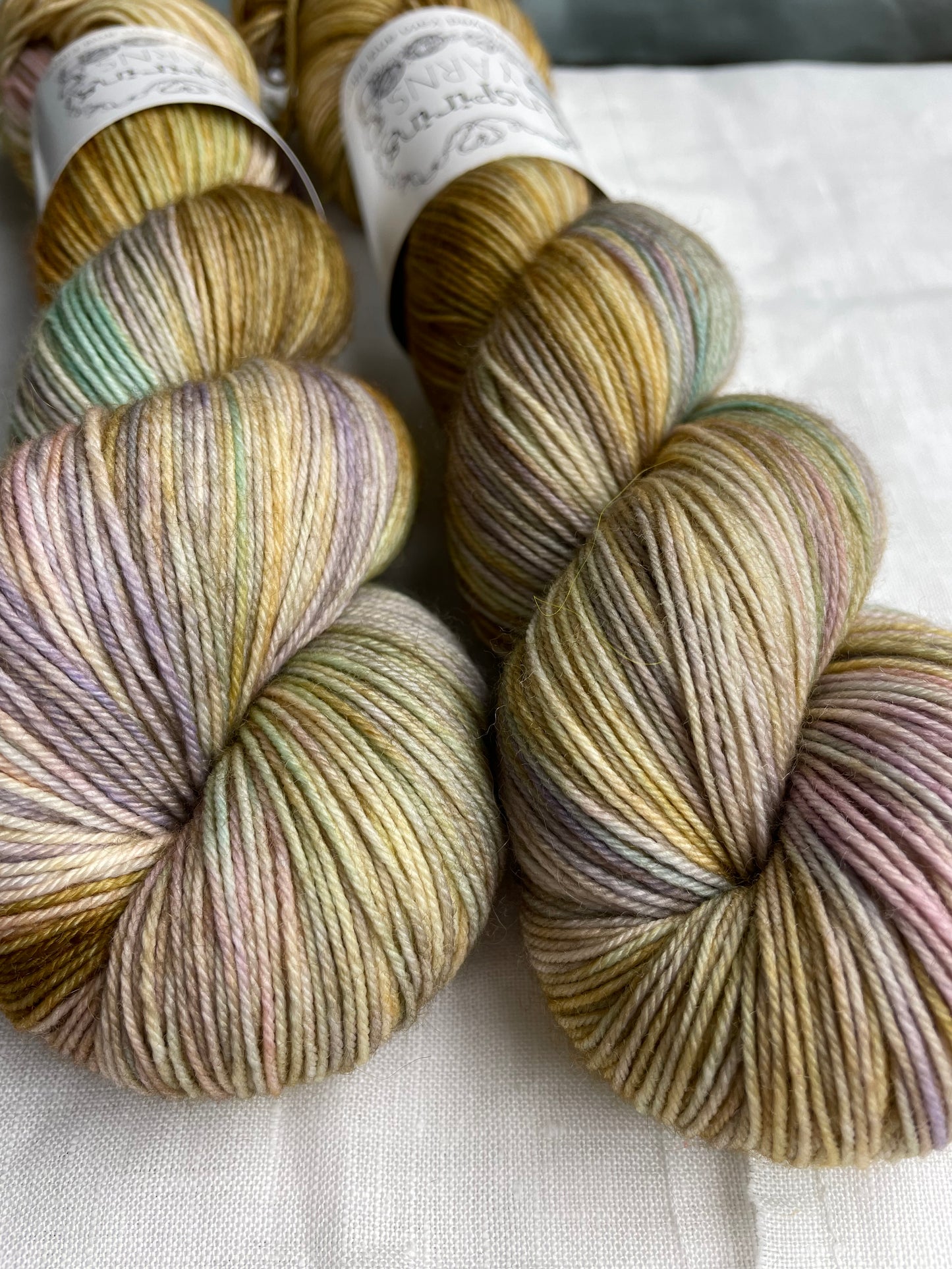 Close up of two soft gold and mixed pastel coloured skeins of Merino Smooth Sock yarn branded Inspiring Yarns on a white linen fabric background.