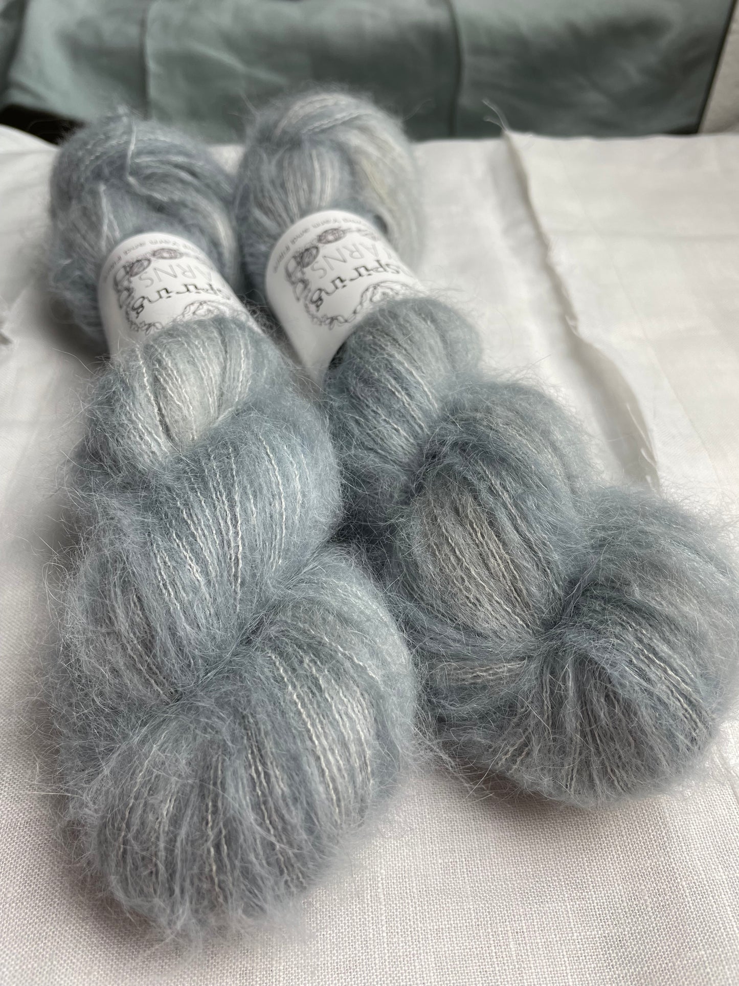Two skeins of fluffy Brushed Suri Silk Lace yarn hand dyed in a pale blue grey colourway and labelled Inspiring Yarns on a white linen background.
