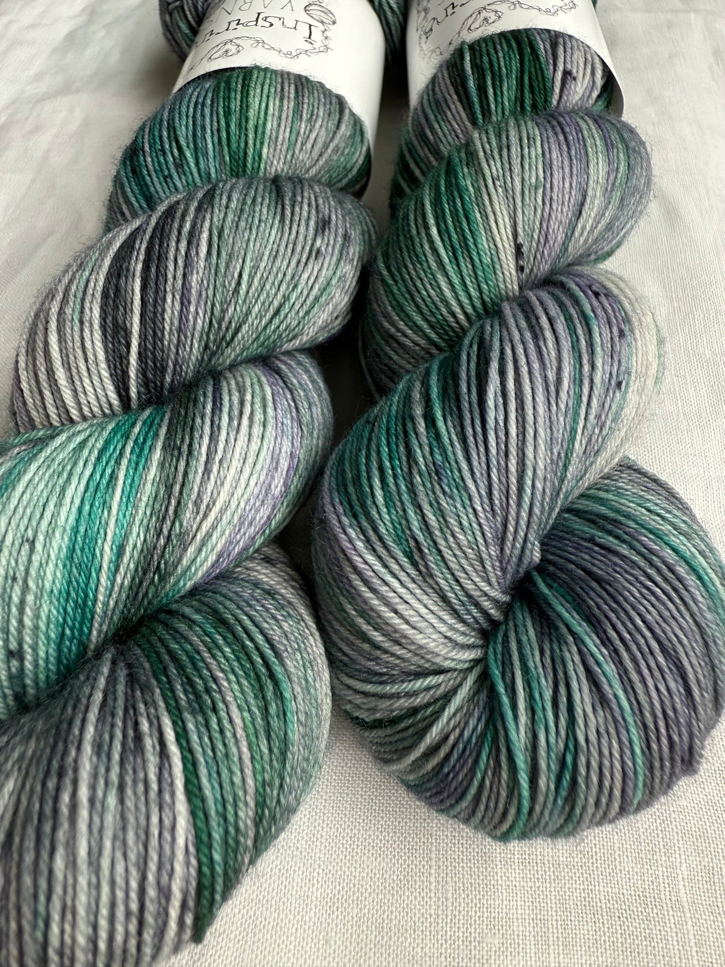 up close image of two skeins of yarn with greys and green with inspiring yarns labels around them and plain white fabric background.