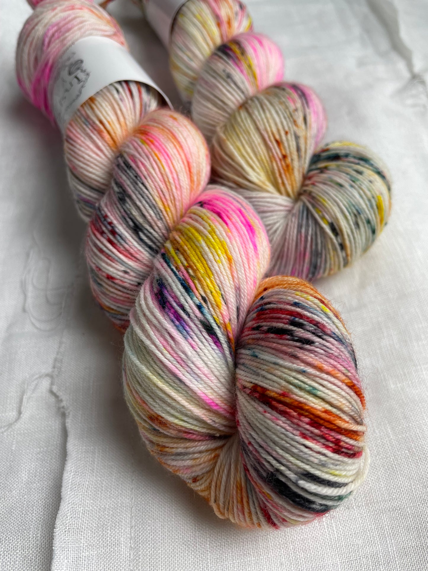 Close up of 2 skeins of multi coloured speckled Merino Smooth Sock labelled InspiringYarns lies on a white linen background.