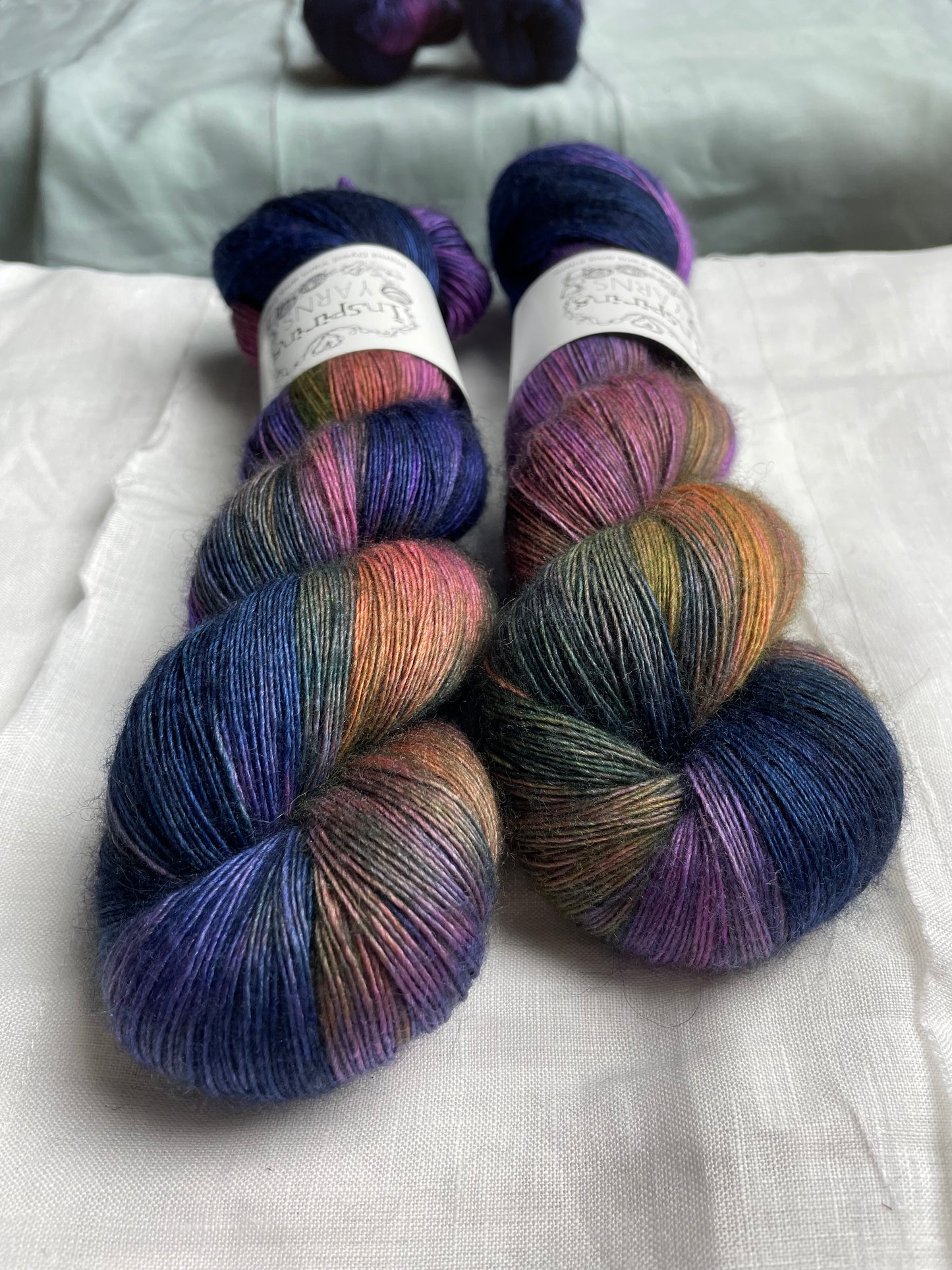 Close up of two variegated multicoloured skeins of hand dyed Yak Silk Singles branded Inspiring Yarns on a white linen background.