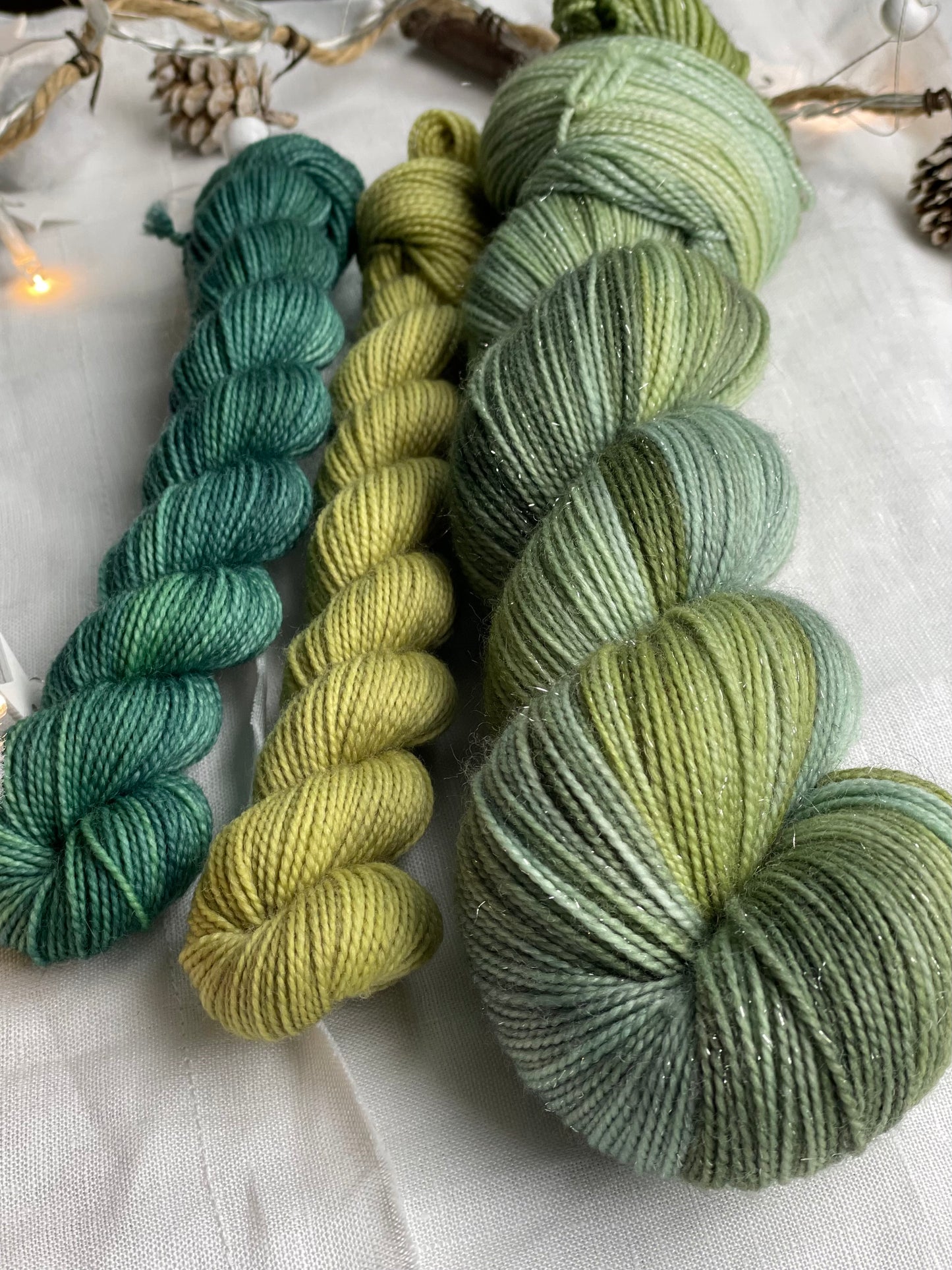 A hand dyed yarn sock set featuring large skein of Sparkle Sock Yarn in green tones with a teal and a lime green mini skein labelled Inspiring Yarns. The background is white linen and some Christmas lights with pinecones, twigs and stars.
