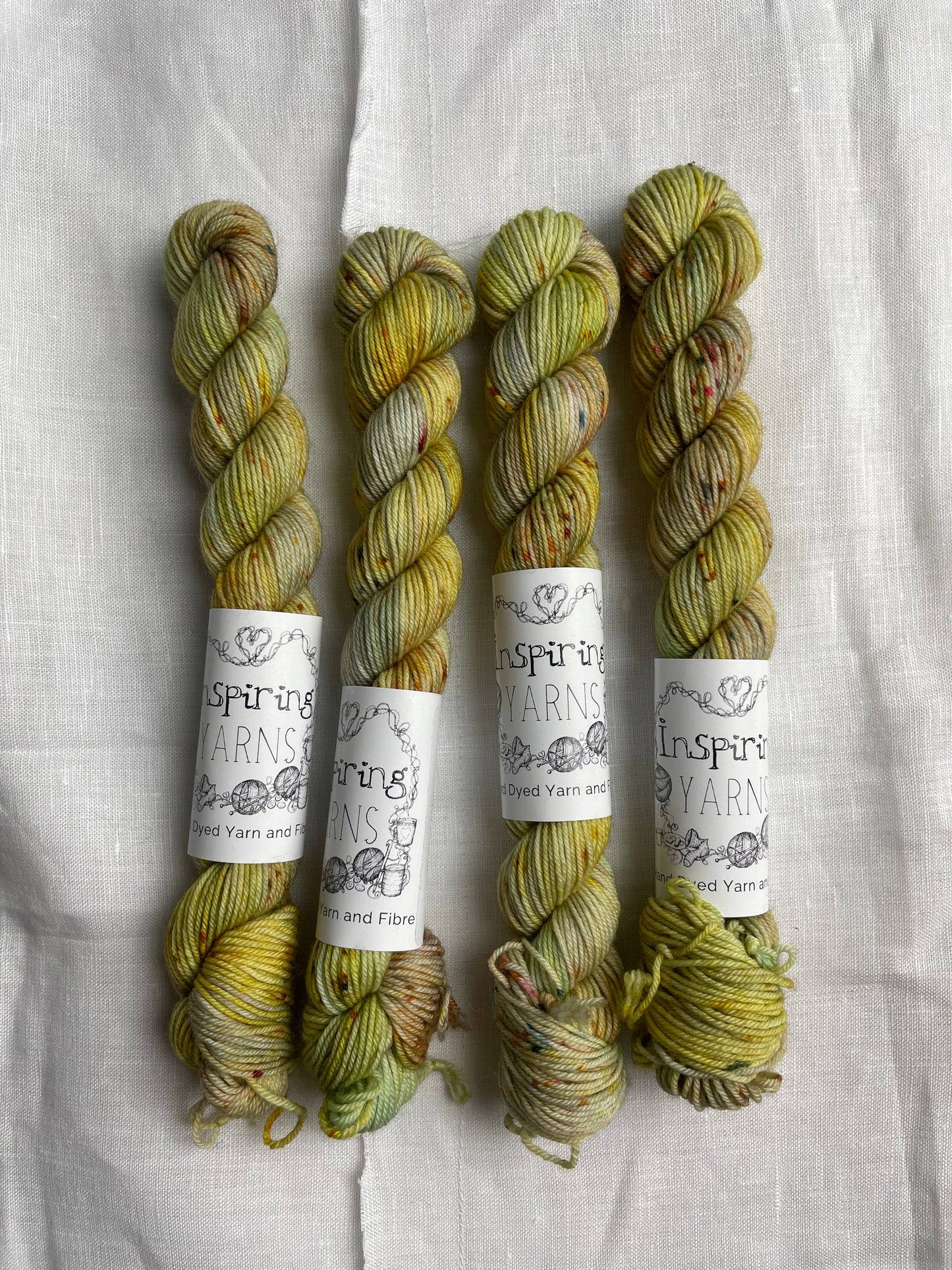 Four yellow based speckled mini skeins of Merino Smooth Sock yarn branded Inspiring Yarns on a white linen fabric background.