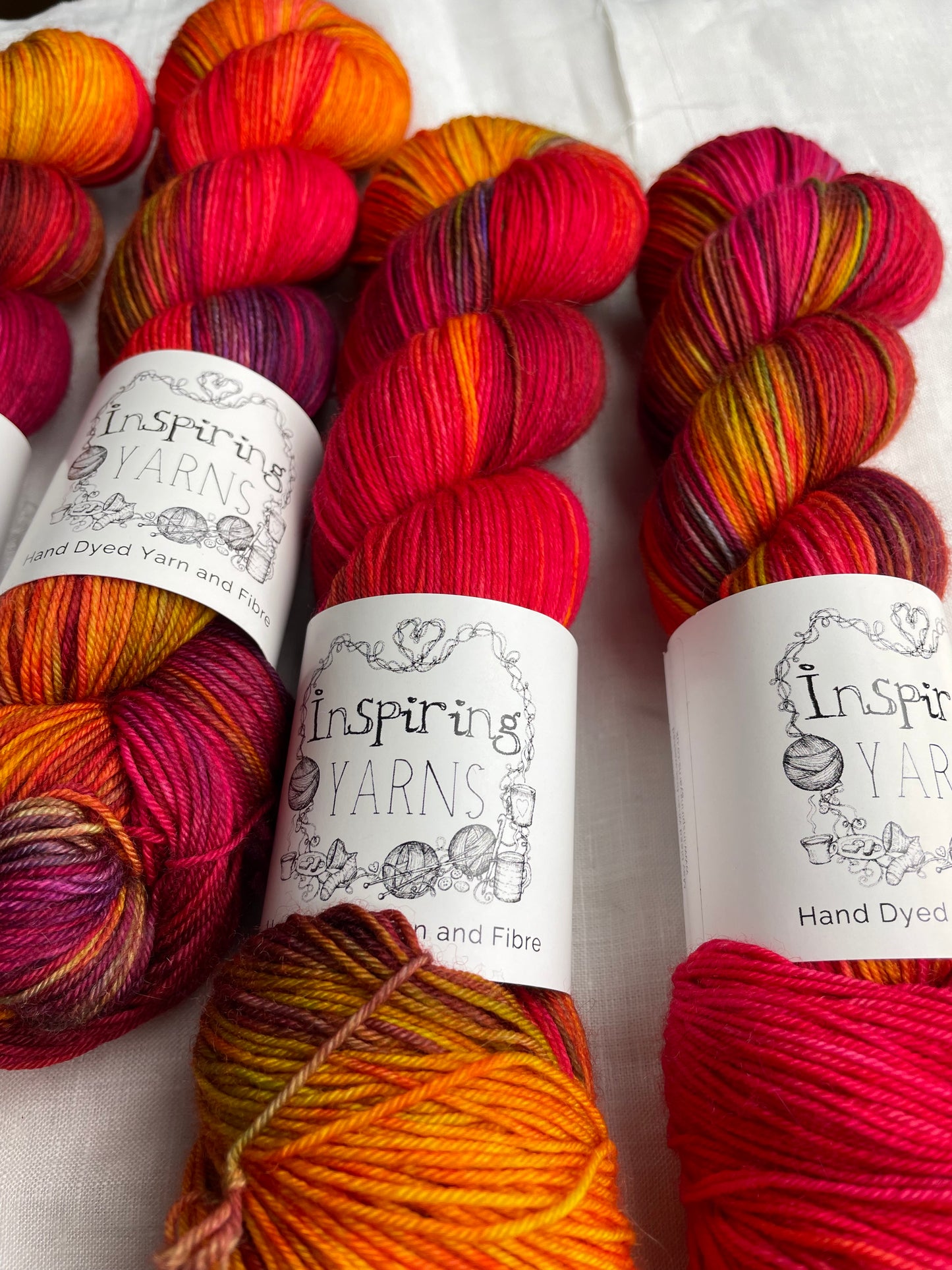 Close up of four variegated red and multi coloured skeins of Merino Smooth Sock yarn branded Inspiring Yarns on a white linen fabric background.