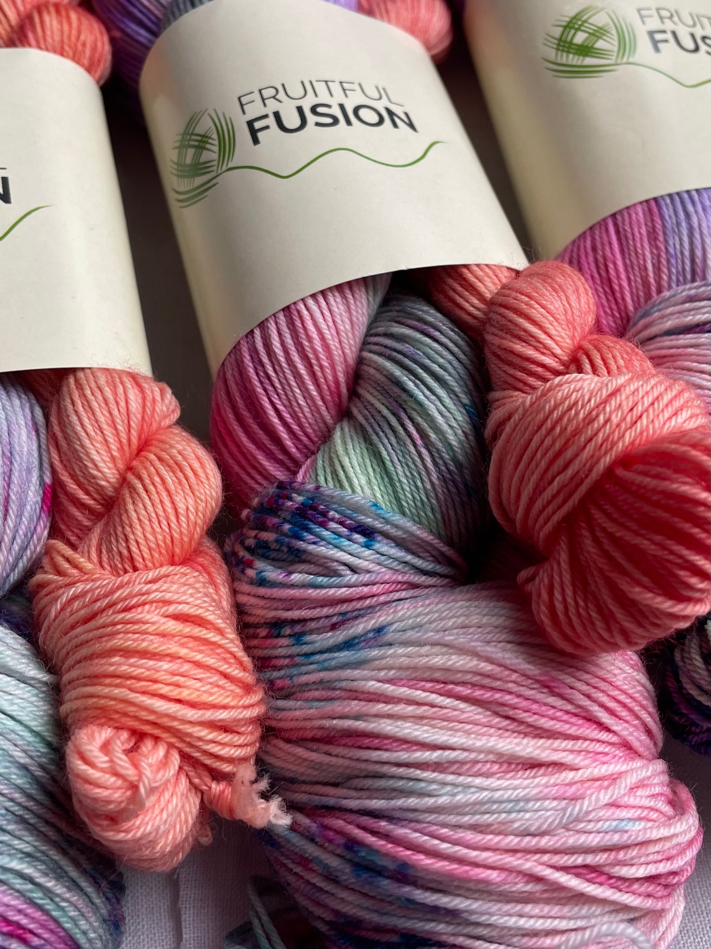 close up of a skein hand dyed yarn in a mix of purple, green and pink with speckles alongside a peach mini skein, the yarn label reads fruitful fusion
