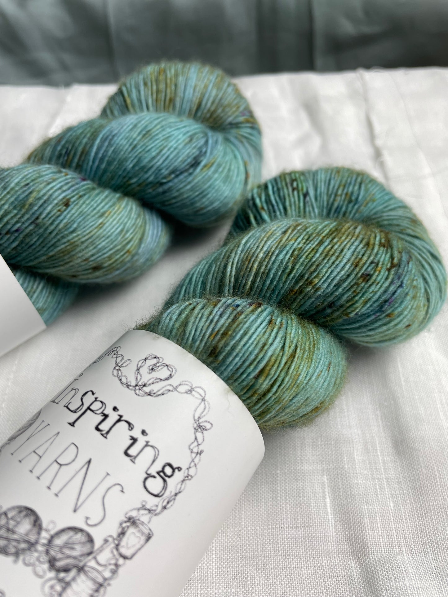 Close up of two skeins of speckled soft teal coloured hand dyed Merino Singles yarn labelled Inspiring Yarns on a white linen background.