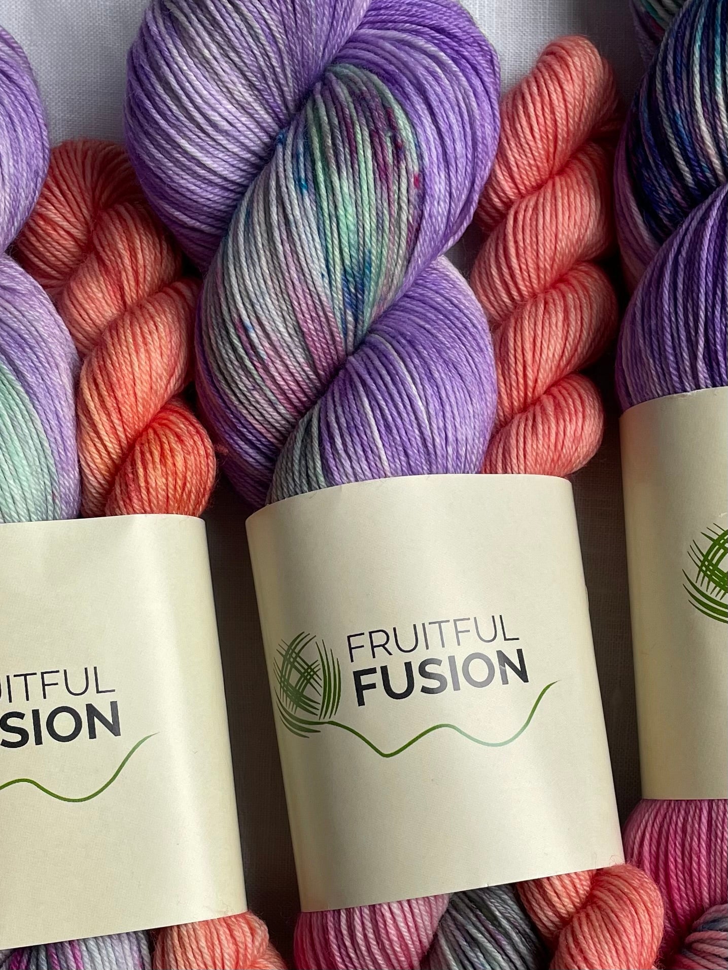 close up of a skein hand dyed yarn in a mix of purple, green and pink with speckles alongside a peach mini skein , the yarn label reads fruitful fusion