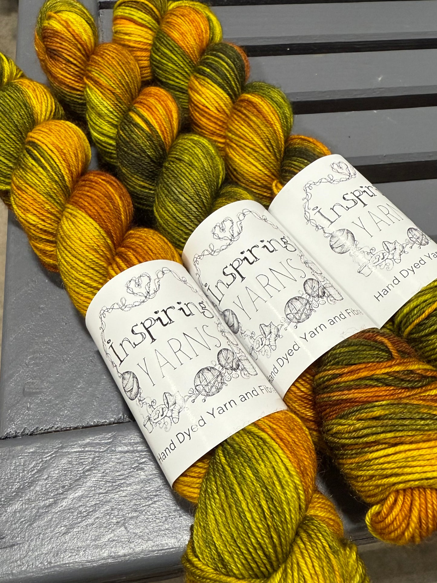 three skeins of yellow green and brown yarn with black and white inspiring yarns labels on a grey wood background.