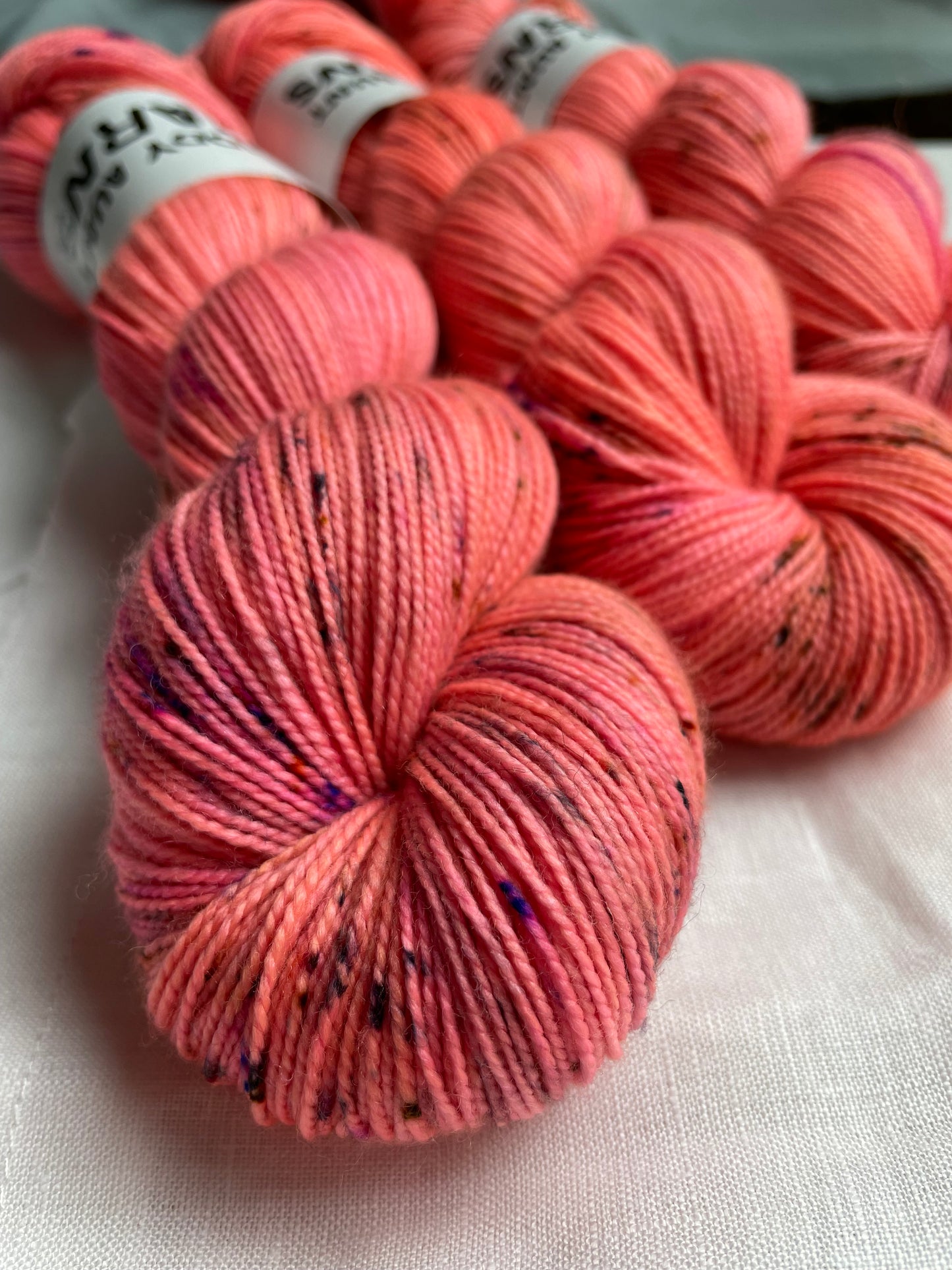close up of 3 skeins of hand dyed yarn in a bright neon coral colour with fuchsia and brown speckles on a white table top with a mirror in the background
