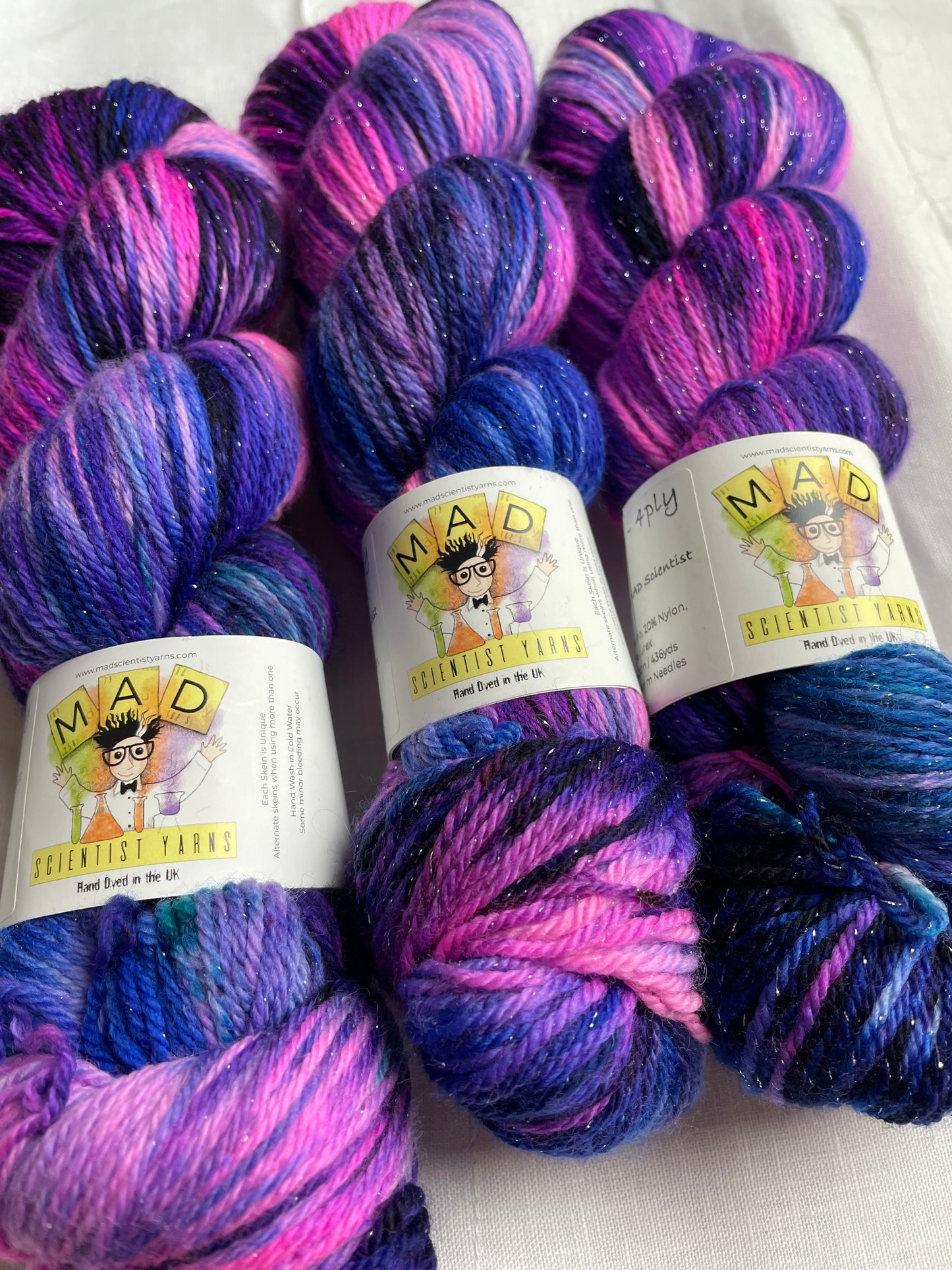 3 skeins of hand dyed yarn in shades of bright pink, purple and blue with silver lurex sparkles lie on a white background, the yarn band reads mad scientist yarns