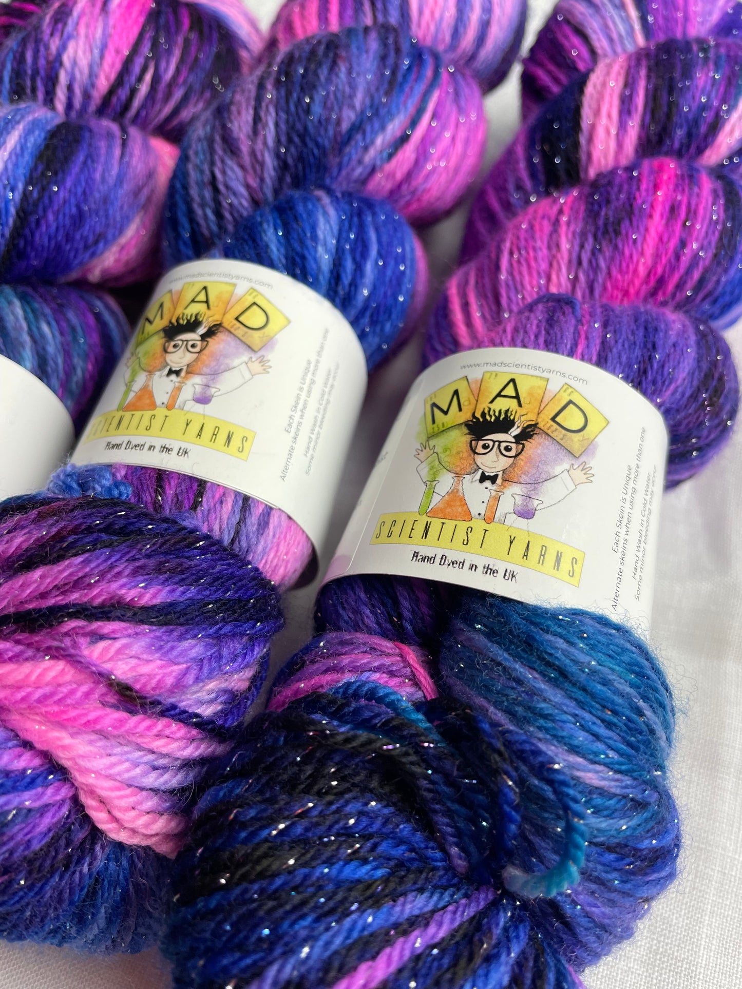 close up of 3 skeins of hand dyed yarn in shades of bright pink, purple and blue with silver lurex sparkles lie on a white background, the yarn band reads mad scientist yarns