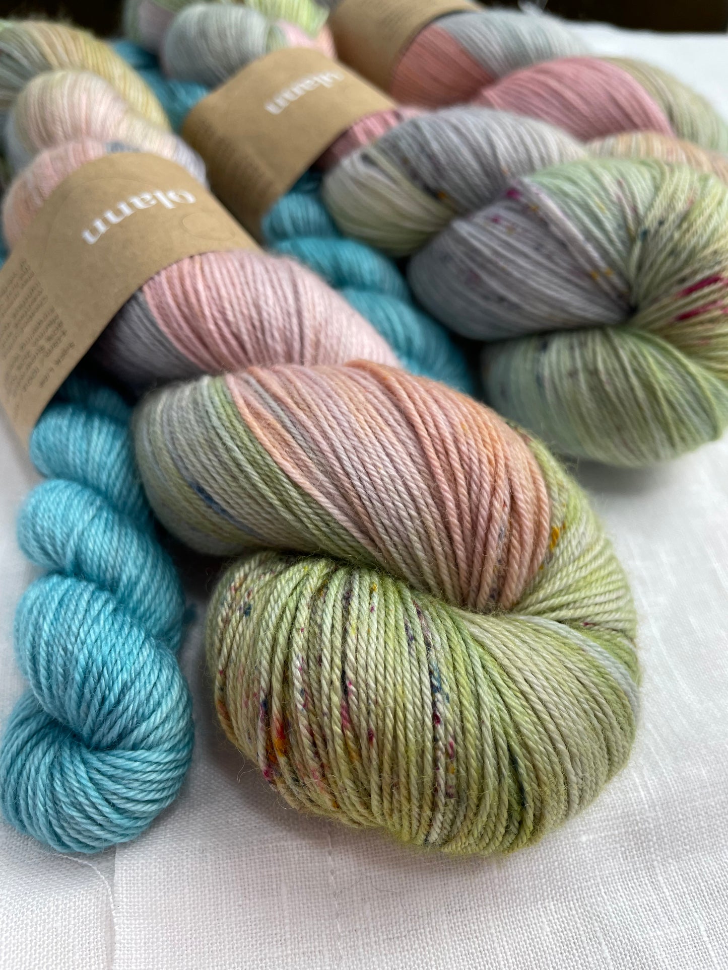 close up of 3 sock sets of hand dyed yarn , the main skein is multi coloured pastel shades and it is paired with a light turquoise mini skein, the yarn is labelled Olann