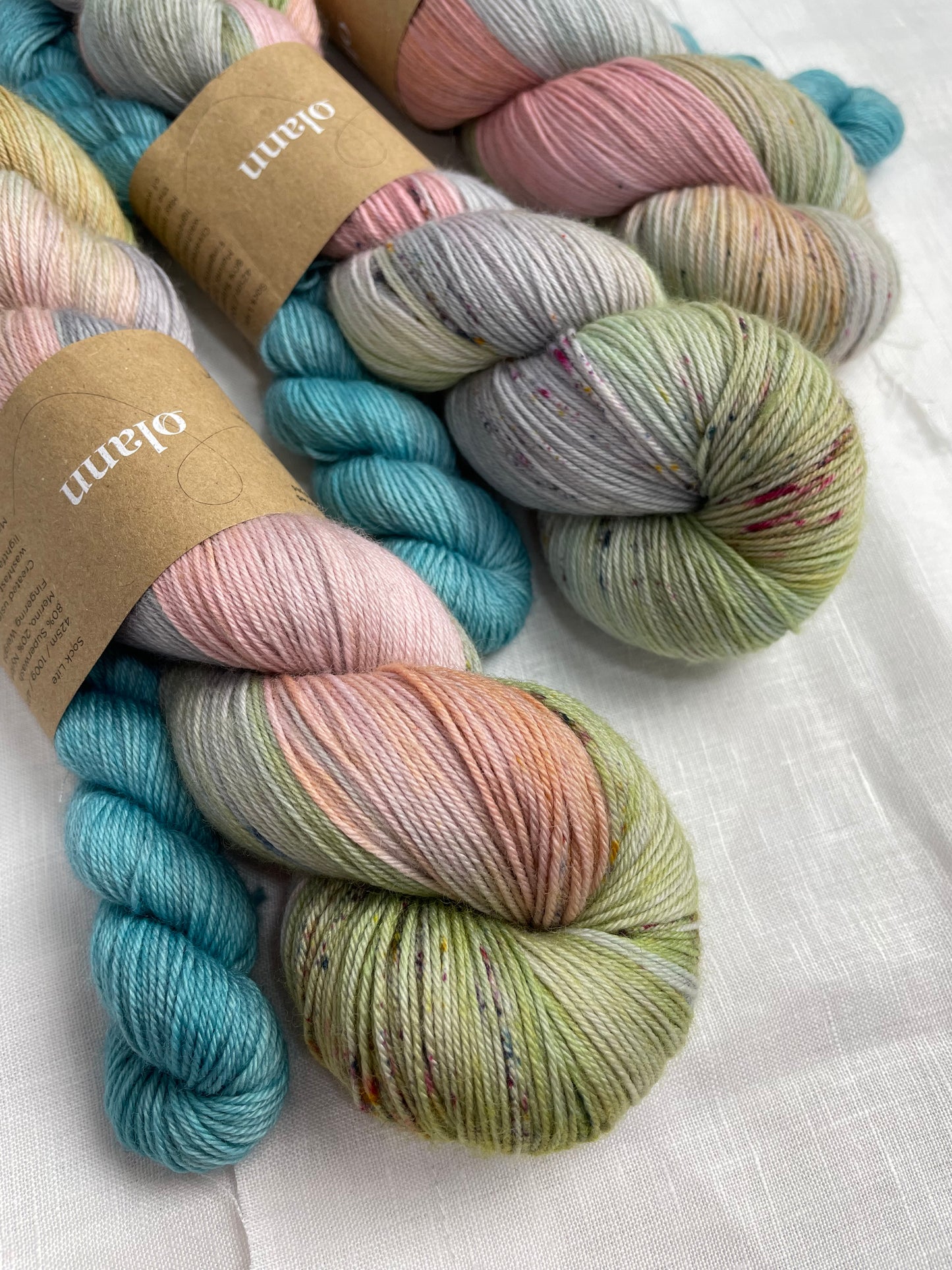 3 sock sets of hand dyed yarn , the main skein is multi coloured pastel shades and it is paired with a light turquoise mini skein, the yarn is labelled Olann on a white background