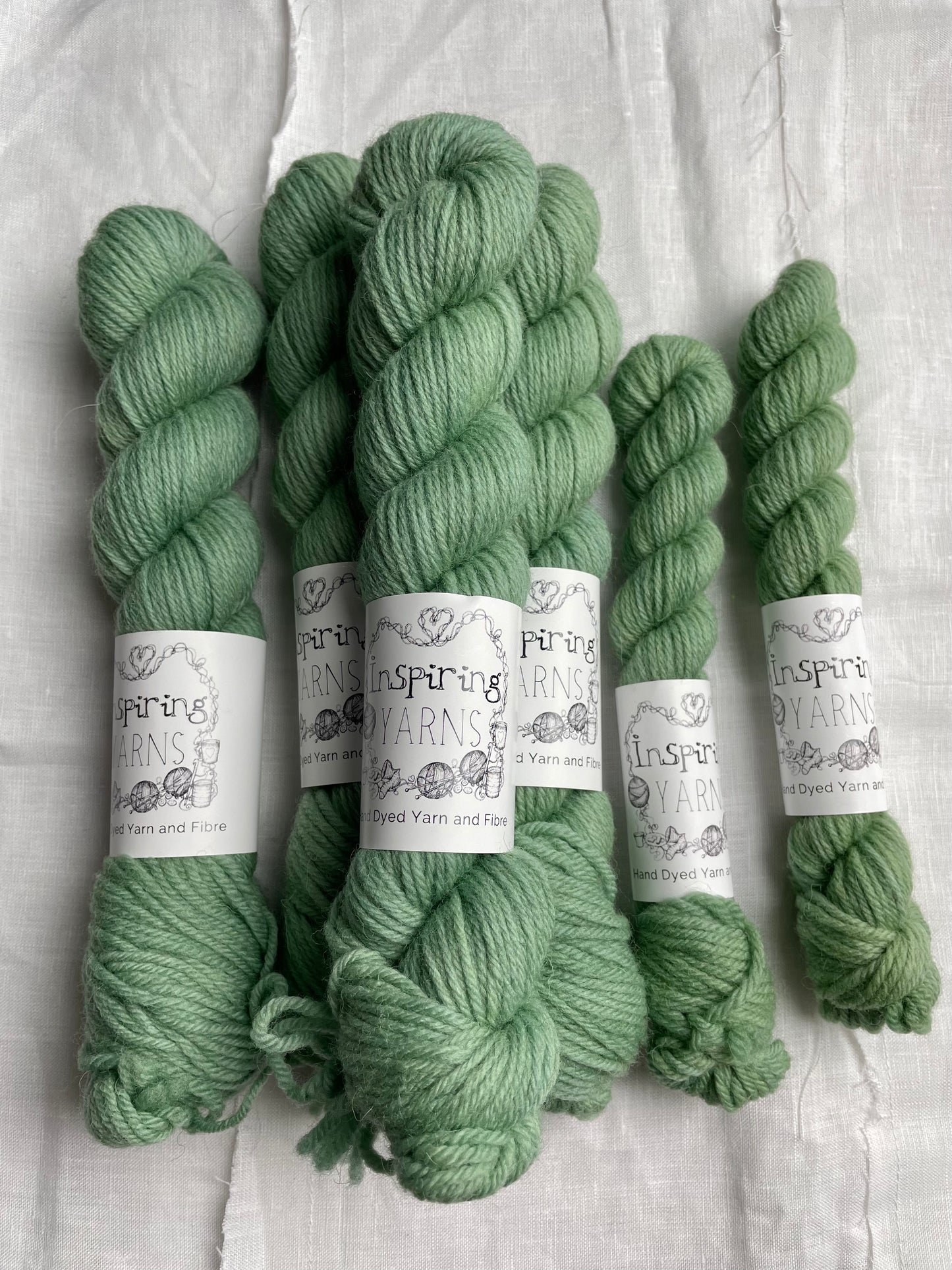 Skeins of mixed sizes of soft mint green coloured Highland DK yarn branded Inspiring Yarns on a white linen background.