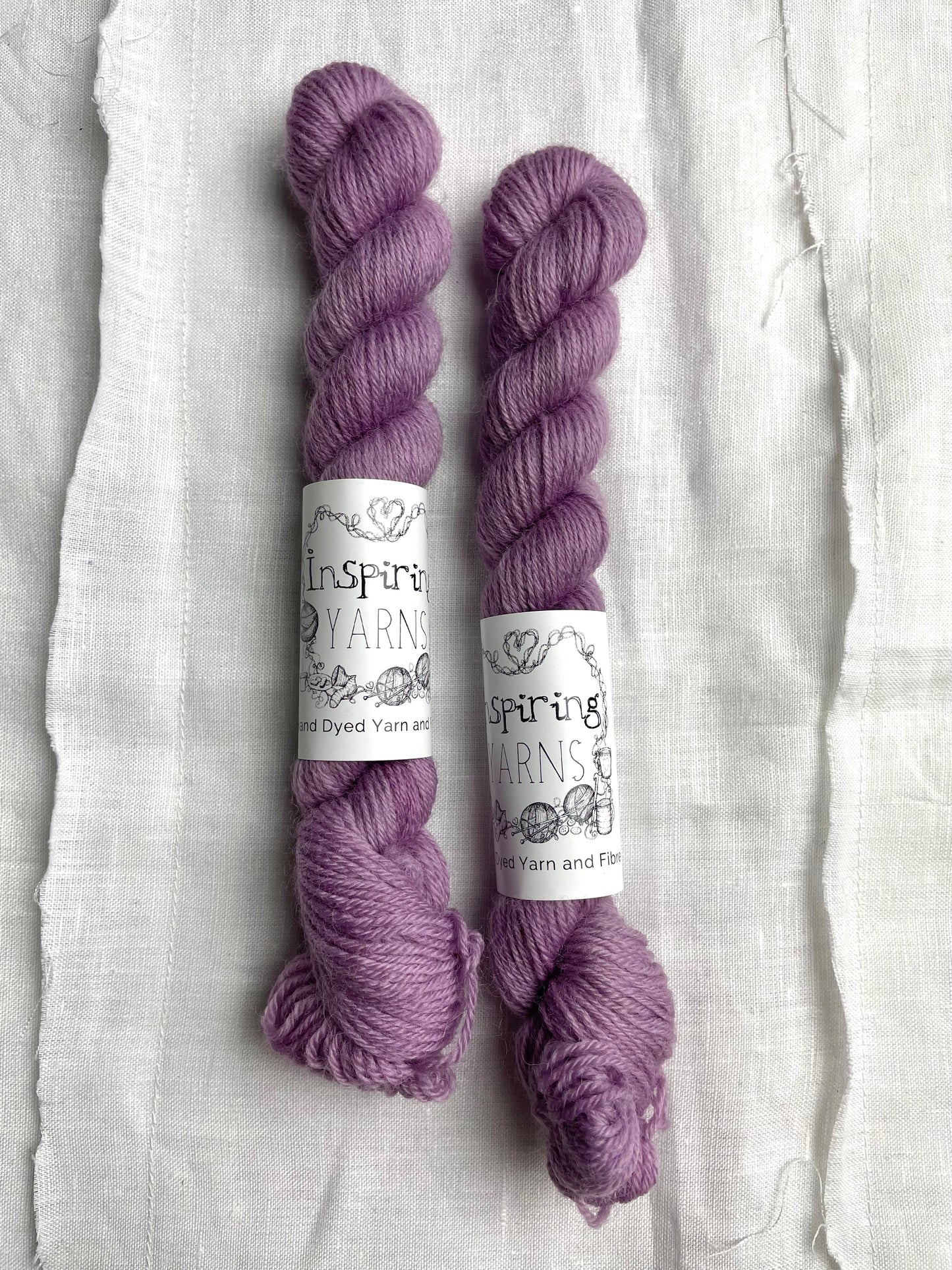 Two mini skeins of tonal dusty lilac coloured Highland 4Ply yarn branded Inspiring Yarns on a white linen background.