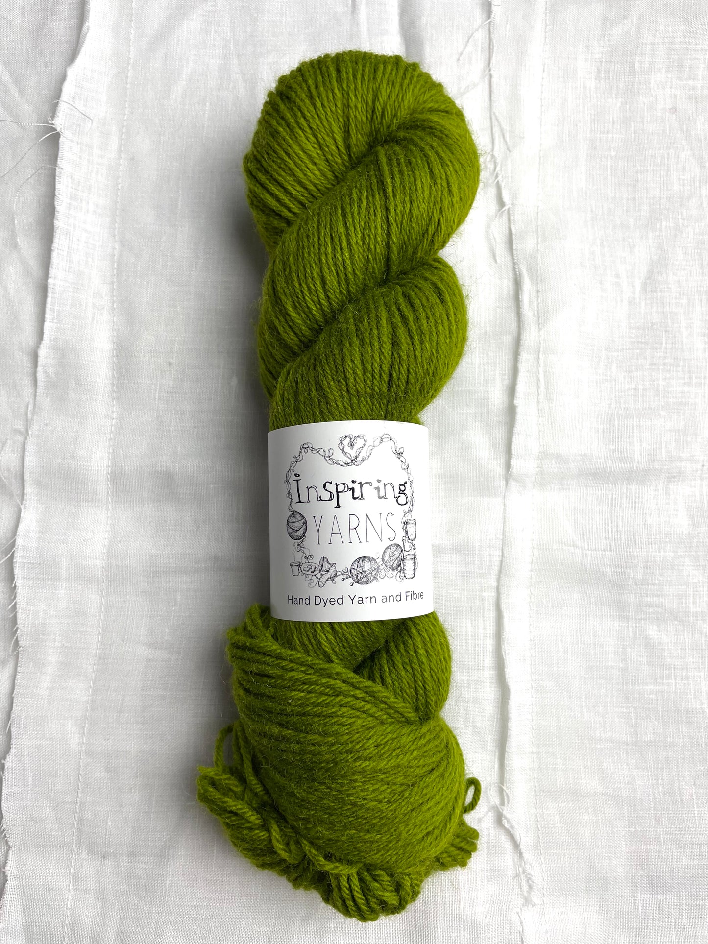 One skein of a tonal dark lime green Highland DK yarn branded Inspiring Yarns on a white linen background.