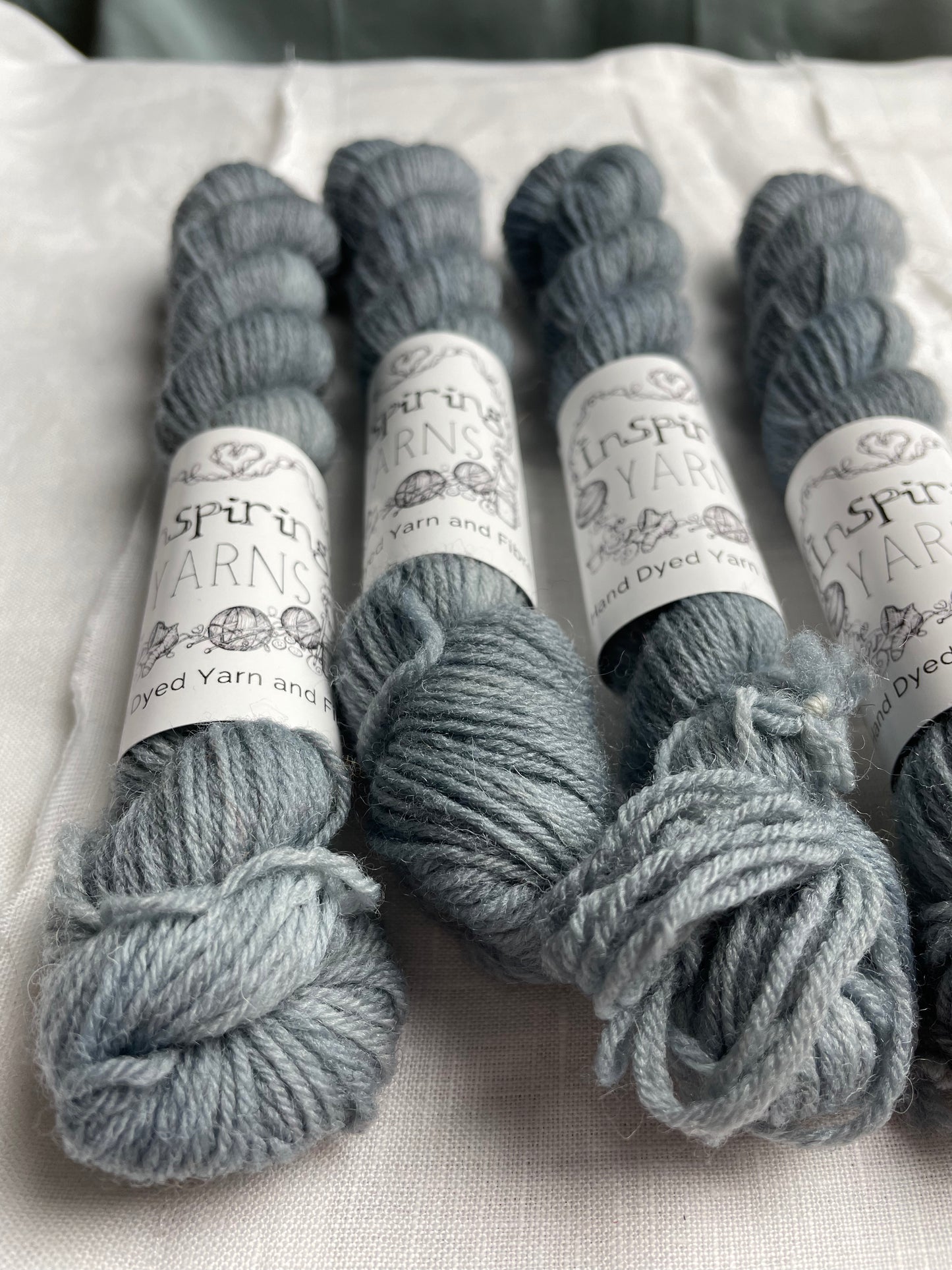 Close up of skeins of tonal light grey blue coloured Highland 4Ply yarn branded Inspiring Yarns on a white linen background.