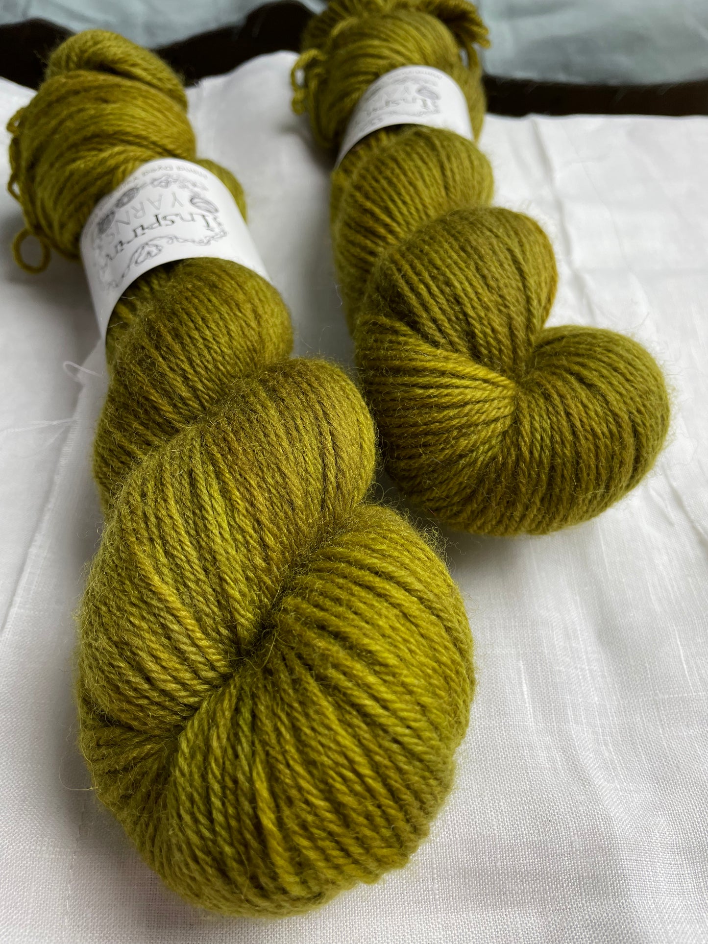 Close up of two skeins of dark olive green coloured Highland DK yarn branded Inspiring Yarns on a white linen background.