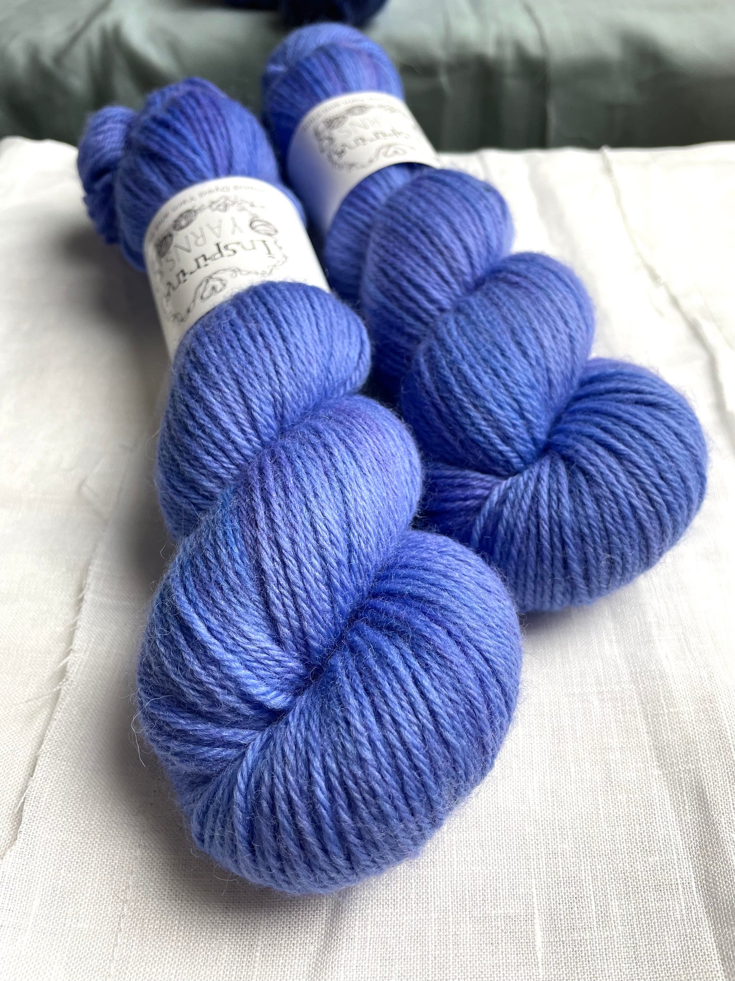 Close up of two skeins of a tonal bright blue lilac Highland DK yarn branded Inspiring Yarns on a white linen background.