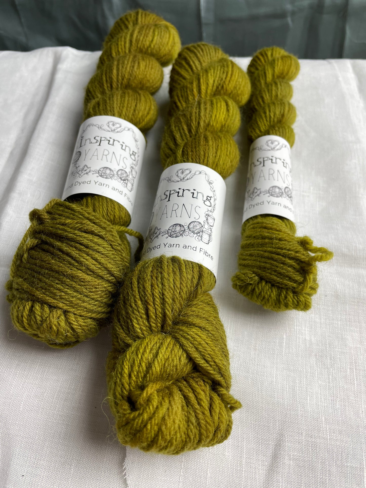 Close up of 3 skeins of dark olive green coloured Highland DK yarn branded Inspiring Yarns on a white linen background.