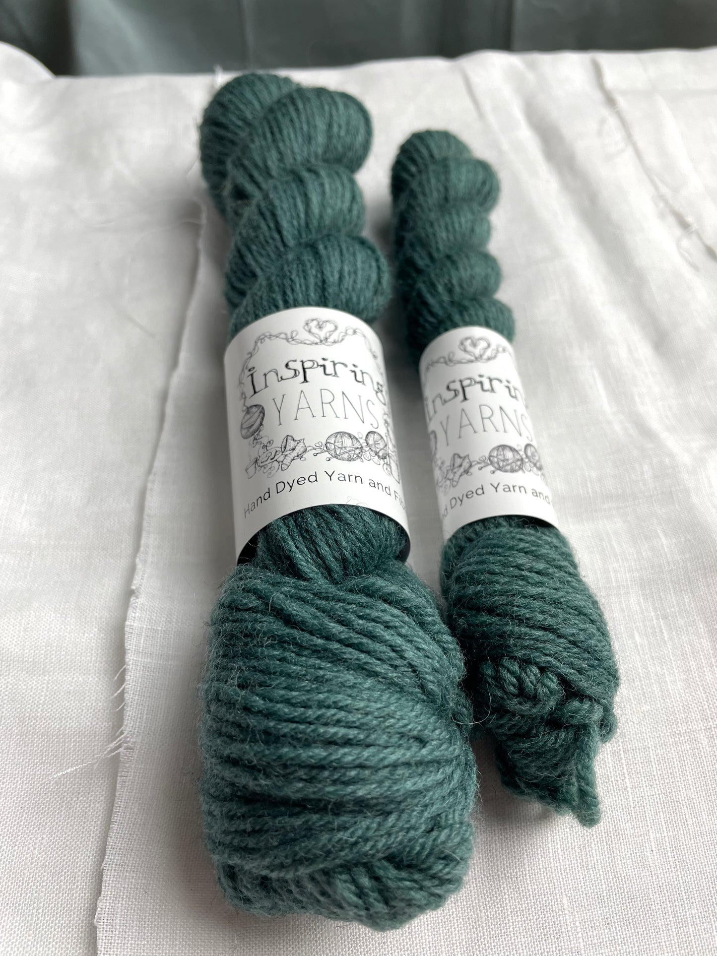Close up of Two skeins of a tonal dark teal Highland DK yarn branded Inspiring Yarns on a white linen background.