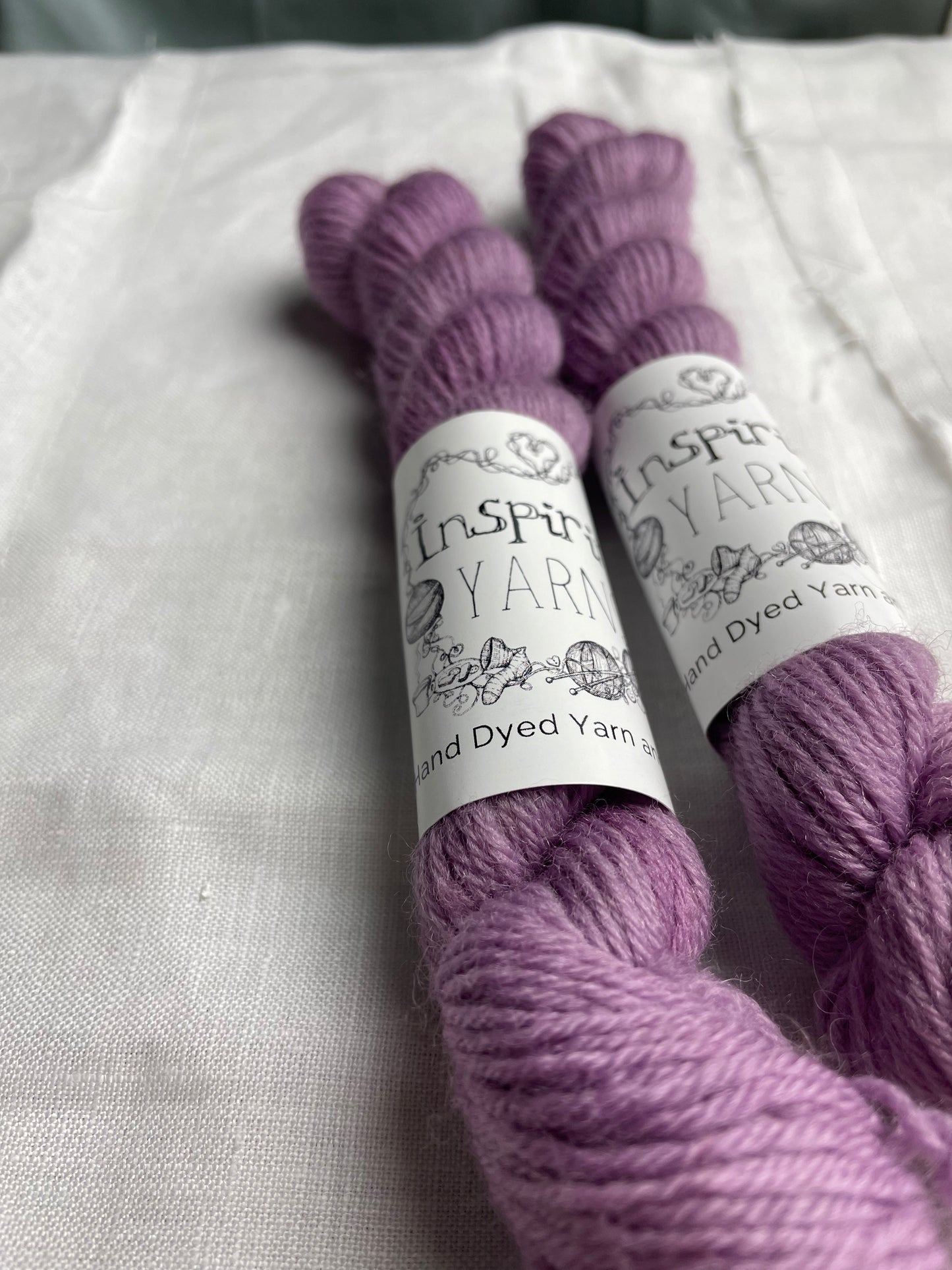 A close up of two mini skeins of tonal dusty lilac coloured Highland 4Ply yarn branded Inspiring Yarns on a white linen background.