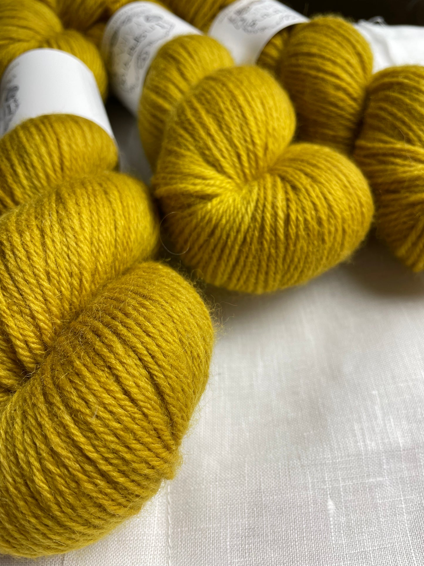 Close up of 3 skeins of tonal golden ochre coloured Highland DK yarn branded Inspiring Yarns on a white linen background.