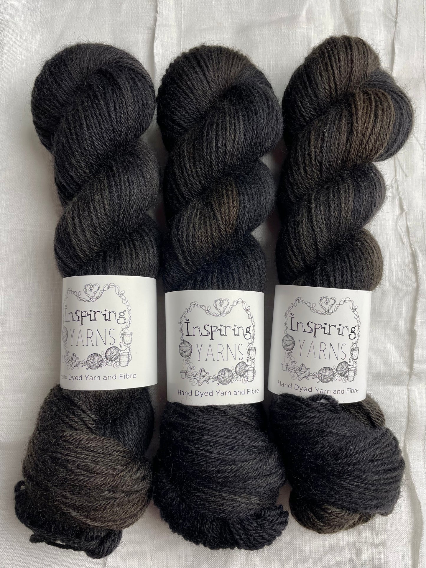3 skeins of tonal black Highland DK yarn lie on a white linen background.