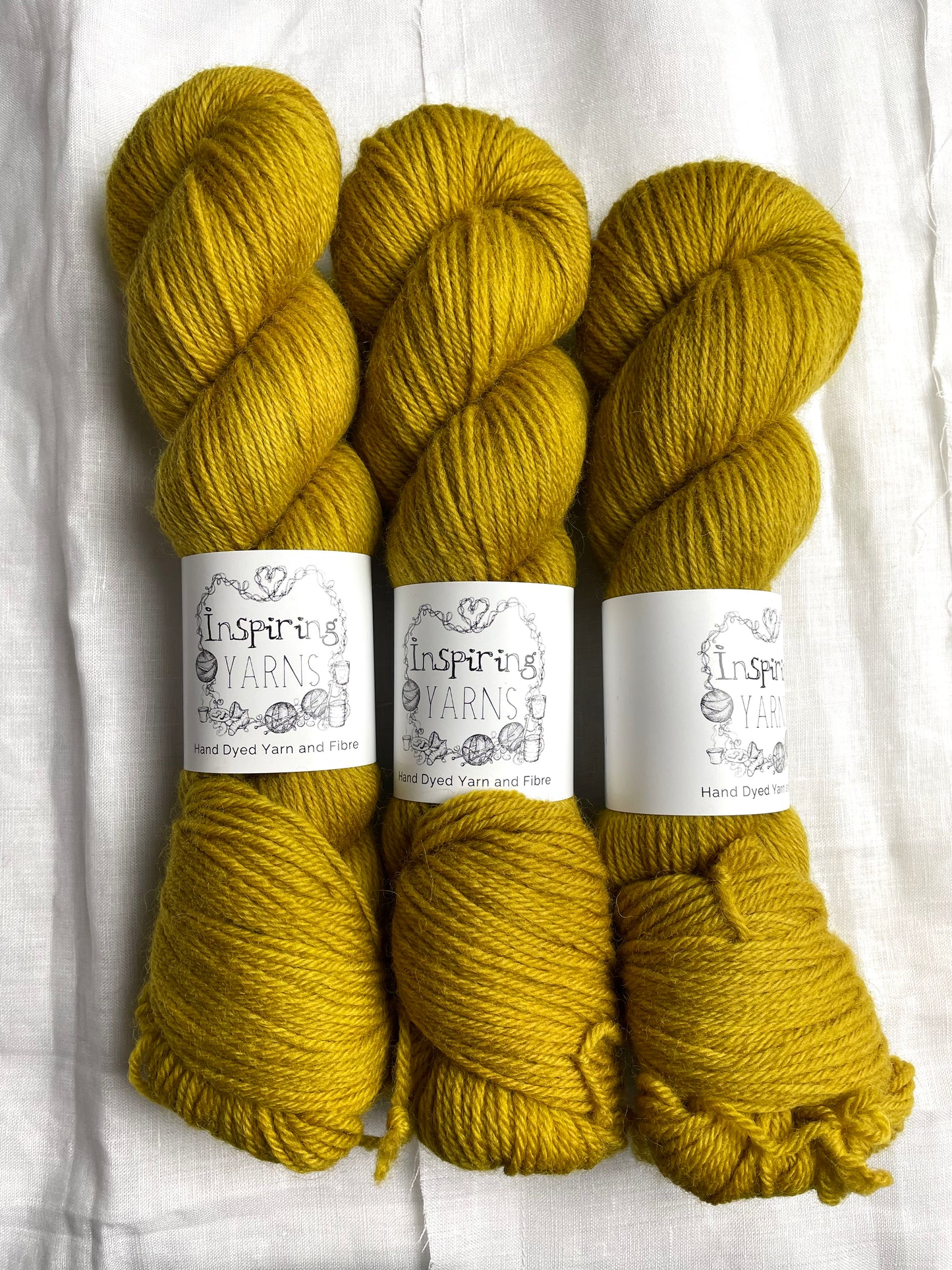 Three skeins of tonal golden ochre coloured Highland DK yarn branded Inspiring Yarns on a white linen background.