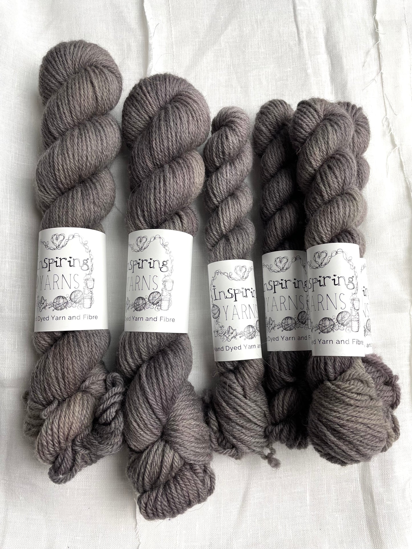 Three skeins of a tonal grey Highland DK yarn branded Inspiring Yarns on a white linen background.
