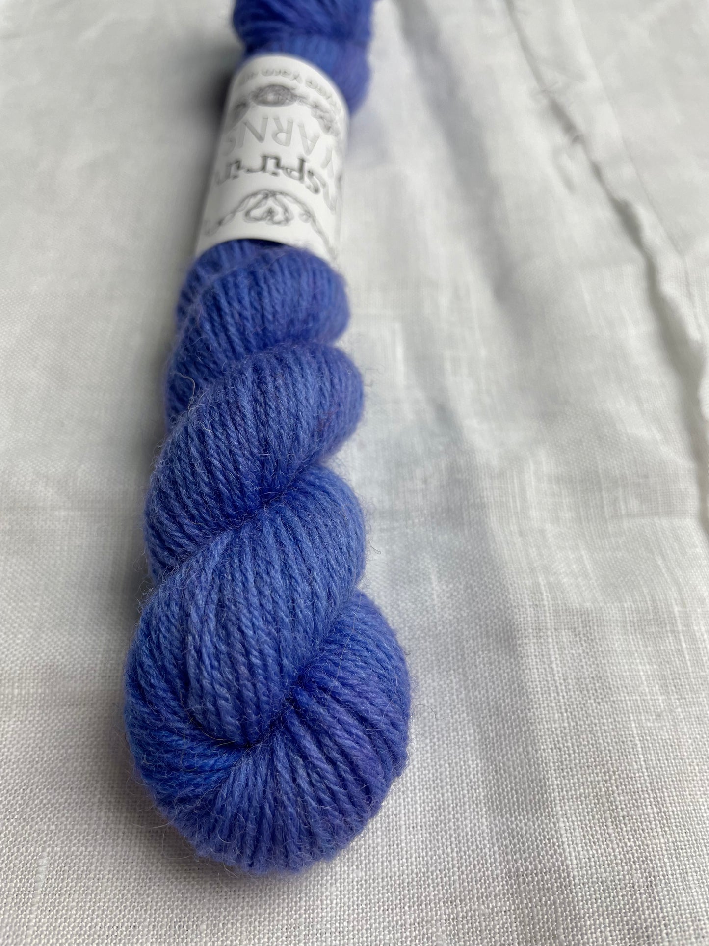 Close up of a mini skein of tonal bright blue lilac coloured Highland 4Ply yarn branded Inspiring Yarns on a white linen background.
