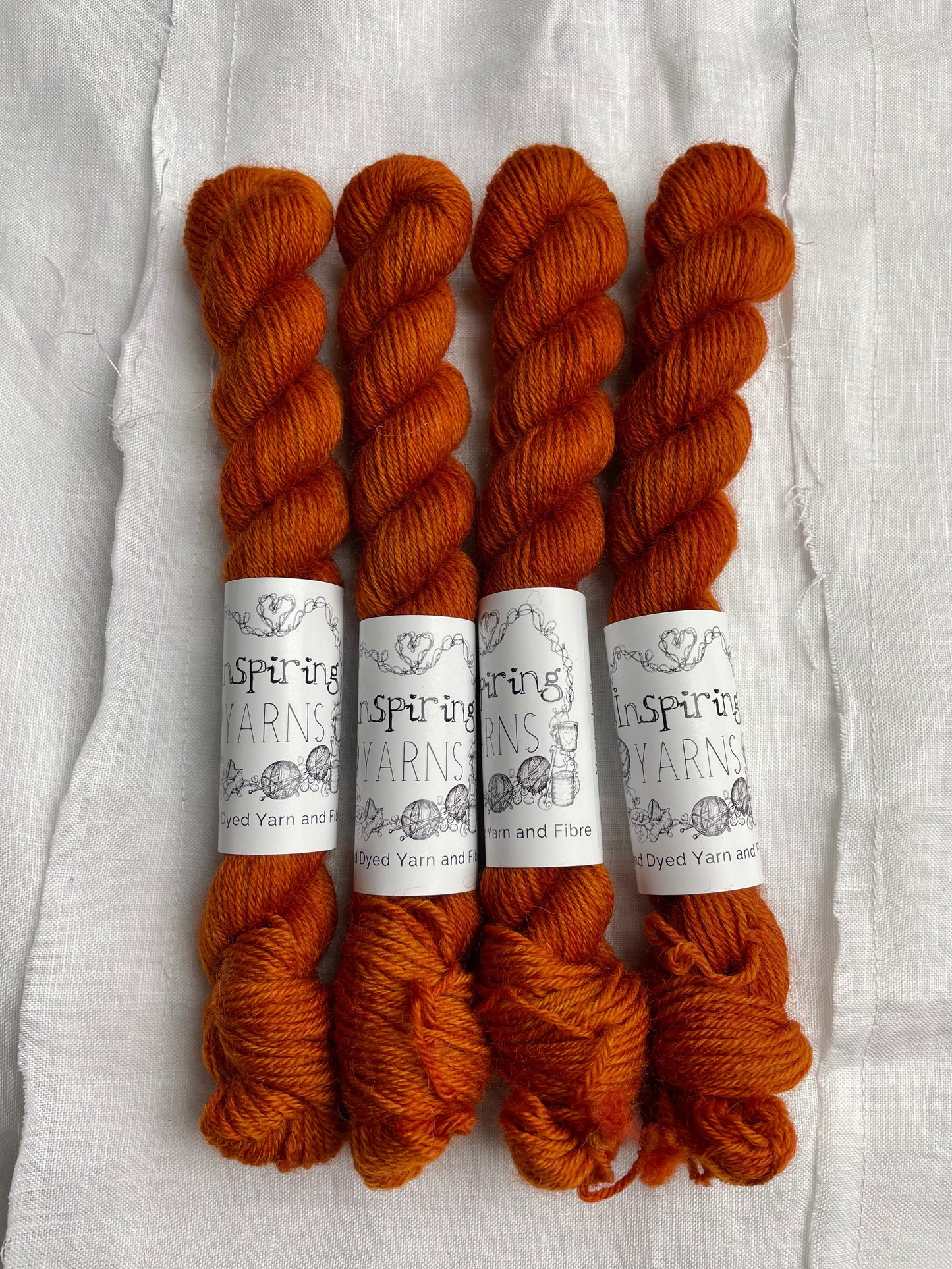 Four mini skeins of tonal burnt orange coloured Highland 4Ply yarn branded Inspiring Yarns on a white linen background.