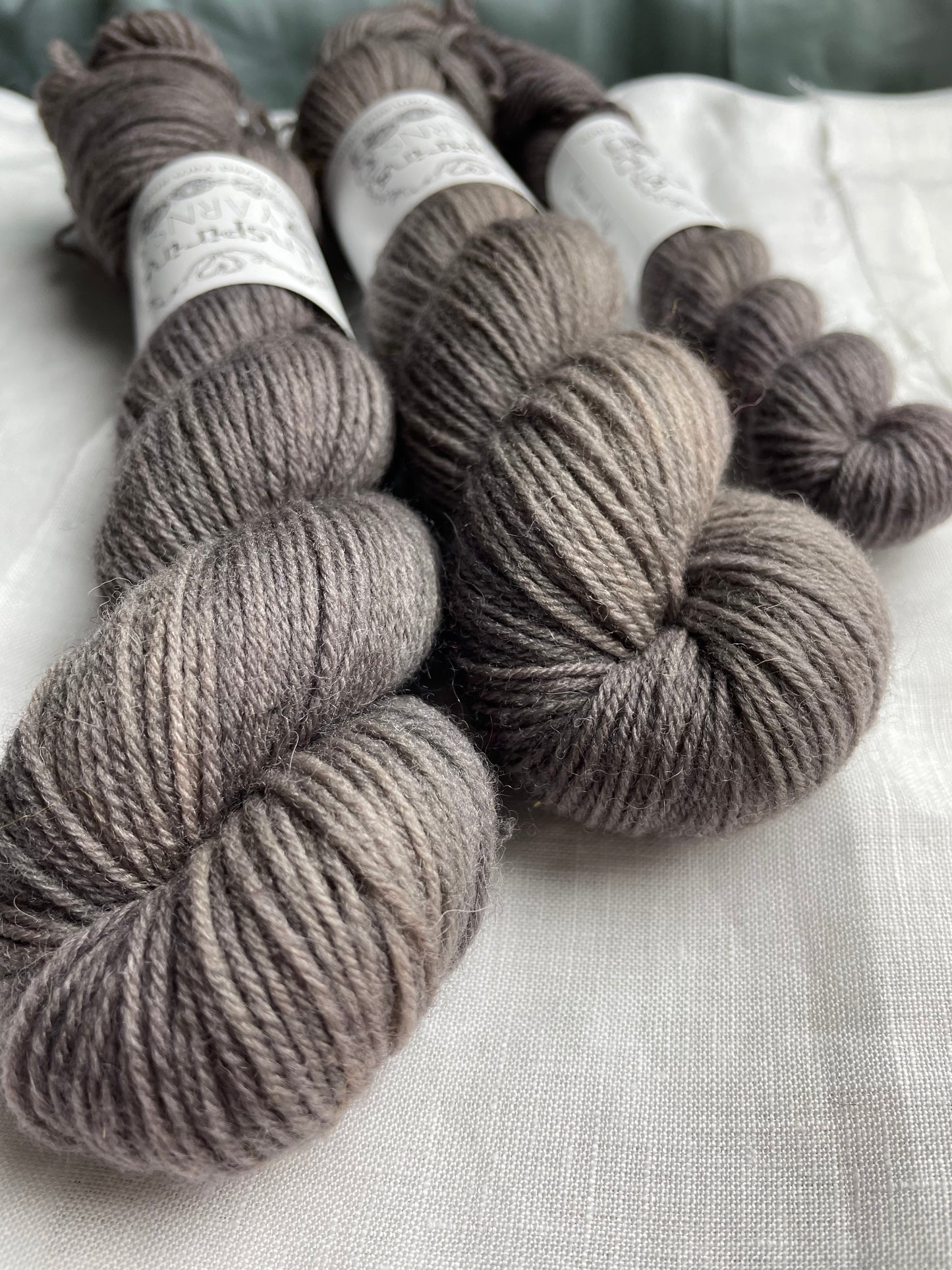 Close up of skeins of tonal dark grey coloured Highland 4Ply yarn branded Inspiring Yarns on a white linen background.