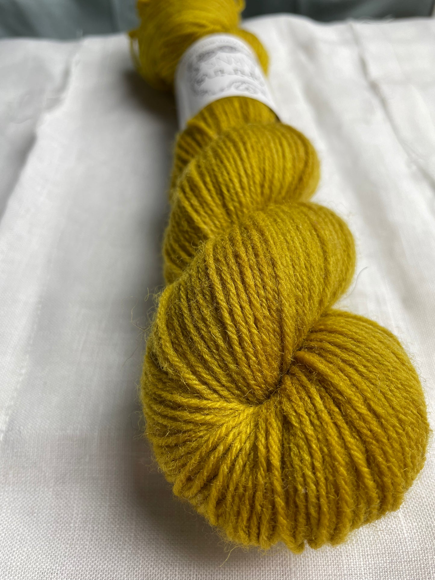Close up of a skein of tonal ochre yellow coloured Highland 4Ply yarn branded Inspiring Yarns on a white linen background.
