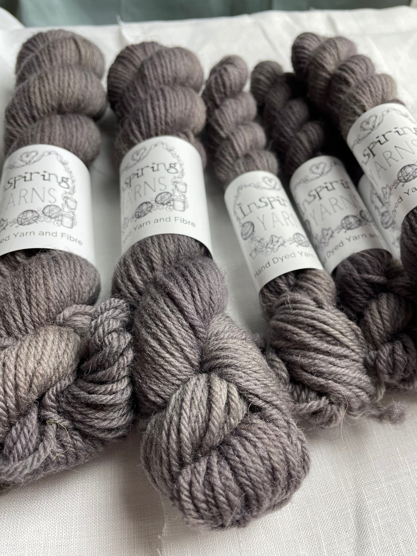Close up of three skeins of a tonal grey Highland DK yarn branded Inspiring Yarns on a white linen background.