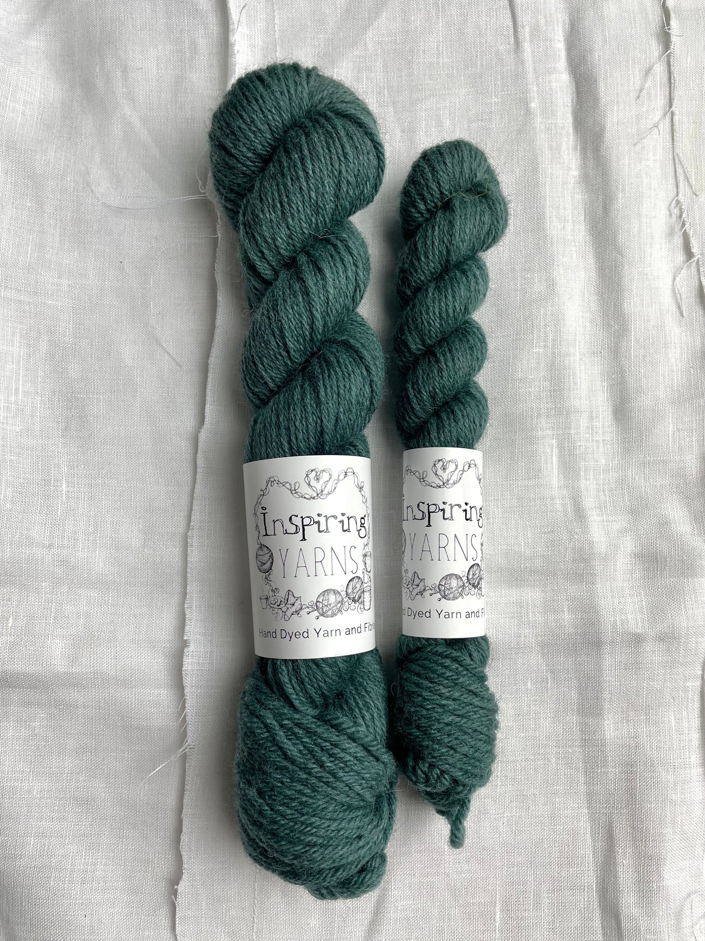 Two skeins of a tonal dark teal Highland DK yarn branded Inspiring Yarns on a white linen background.