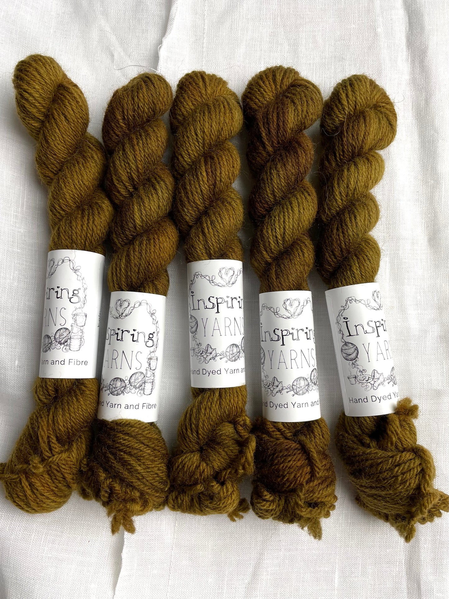 Five mini skeins of a tonal green brown Highland DK yarn branded Inspiring Yarns on a white linen background.