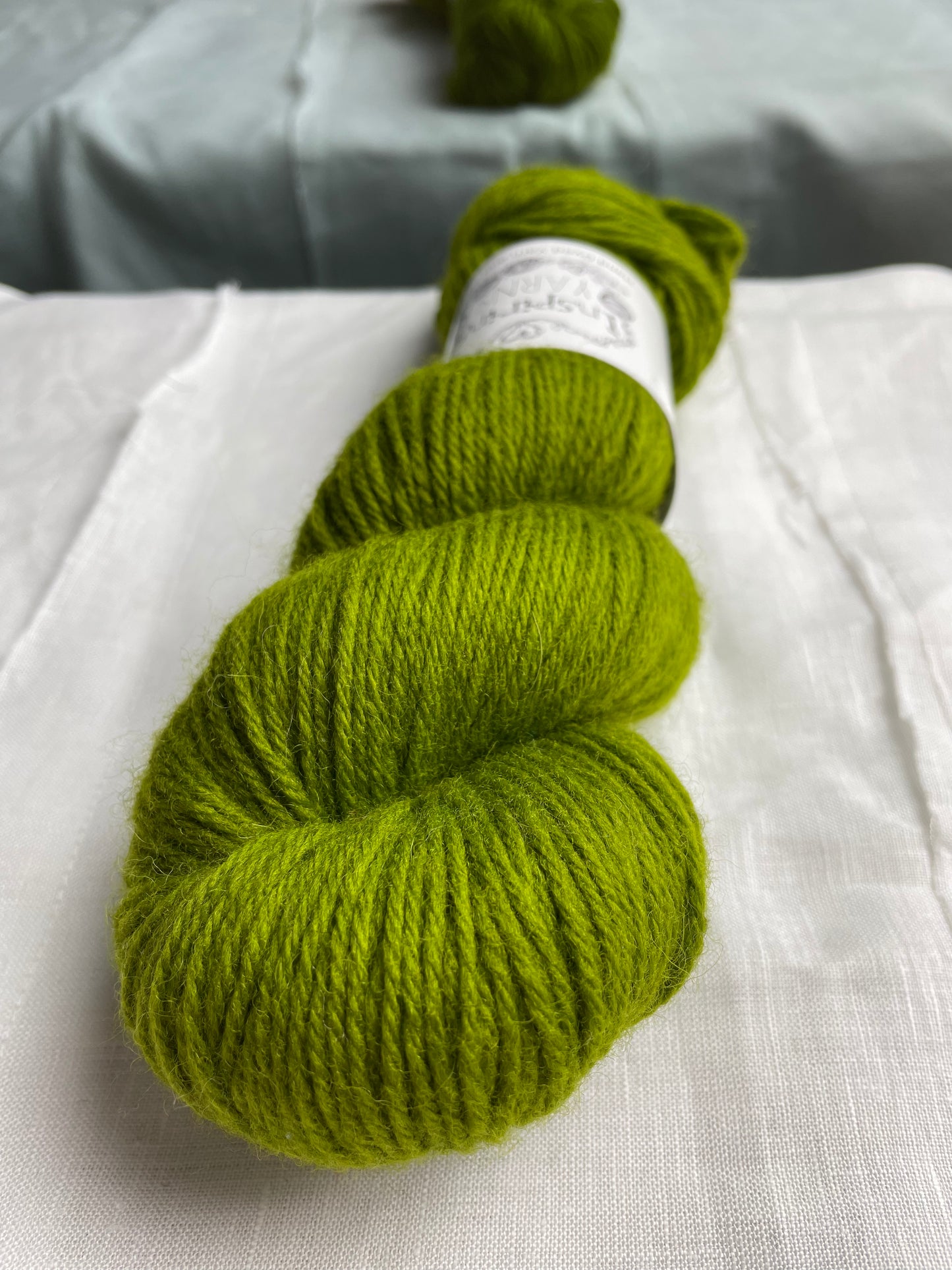 Close up of one skein of a tonal dark lime green Highland DK yarn branded Inspiring Yarns on a white linen background.